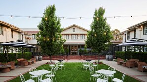 Un patio exterior con mesas y sillas blancas, rodeado de setos y árboles, y luces colgantes.