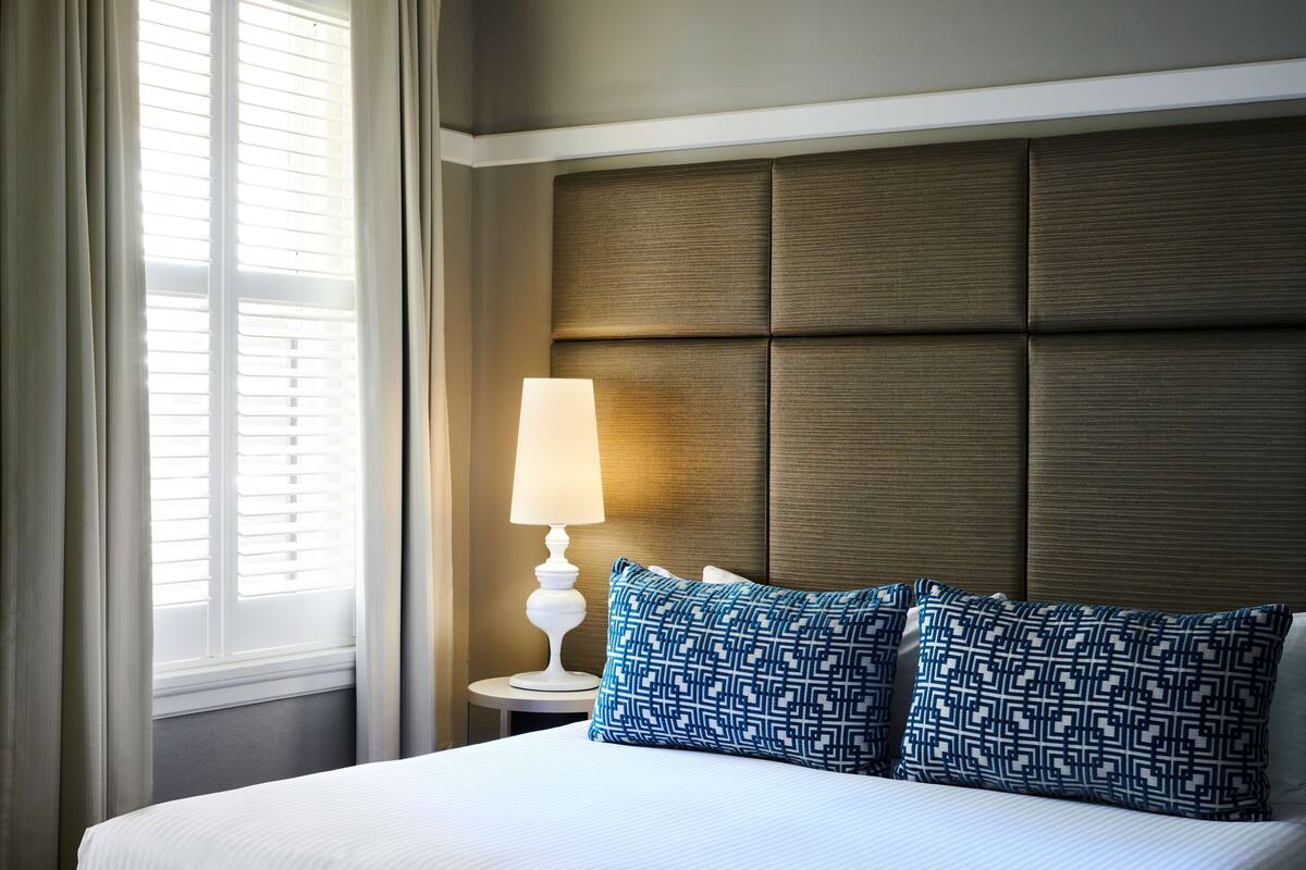 A neatly made bed with a headboard, a bedside lamp, and a window with curtains.