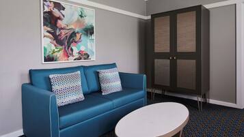 Ein Zimmer mit einem blauen Sofa, zwei gemusterten Kissen, einem weißen Tisch und einem großen, abstrakten Gemälde an der Wand.