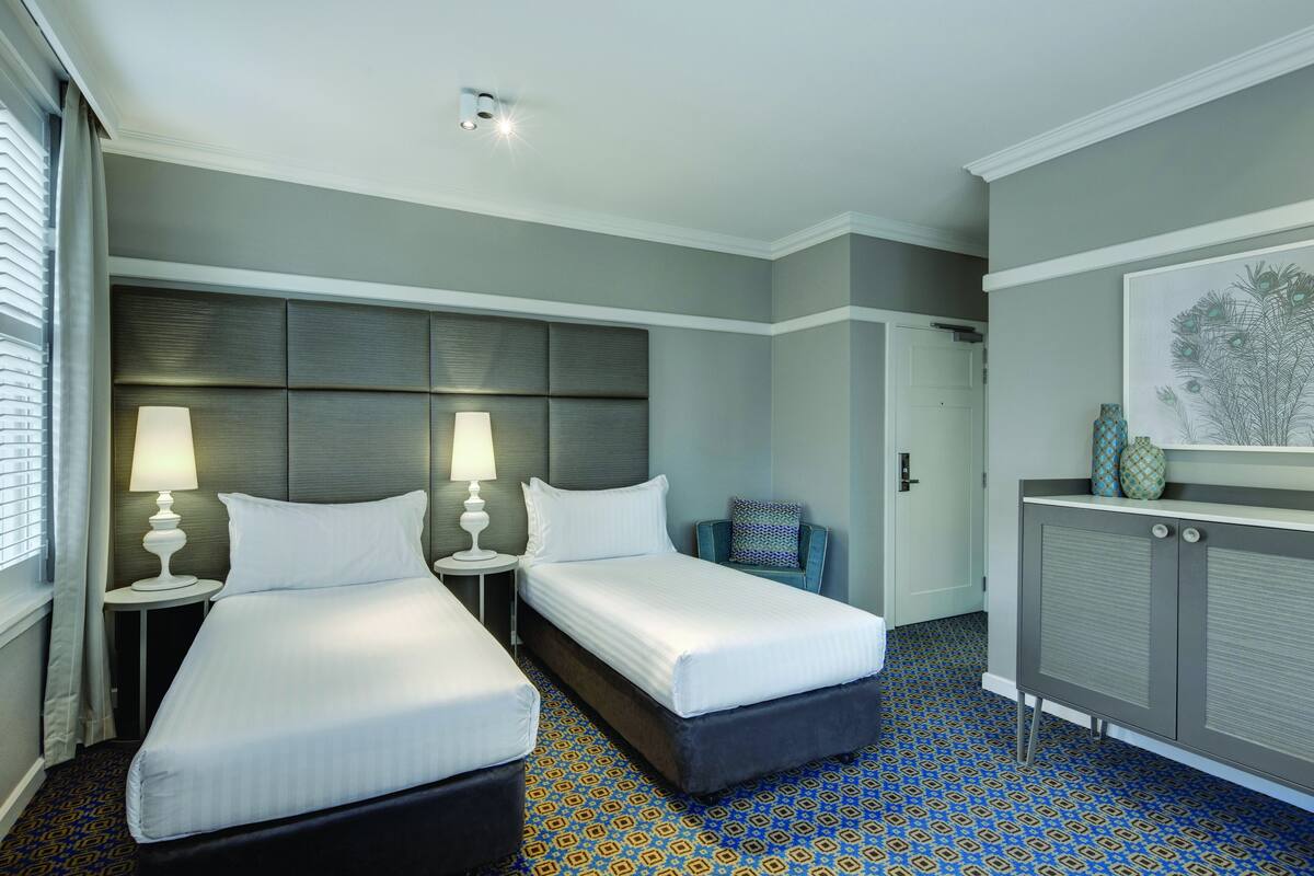 A hotel room with two beds, a nightstand with lamps, a blue patterned carpet, a grey headboard, and a cabinet with decorative items.