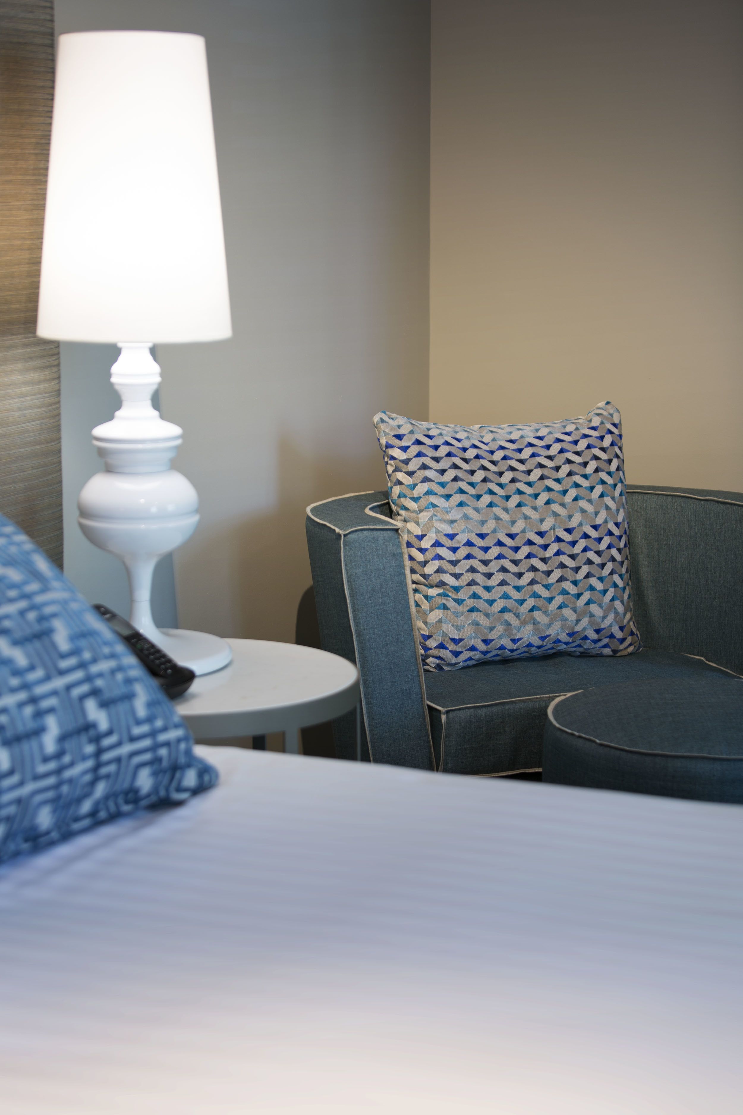 Une chambre d’hôtel avec un lit, une table ronde avec une lampe, une chaise avec un coussin à motifs et une télécommande sur la table.
