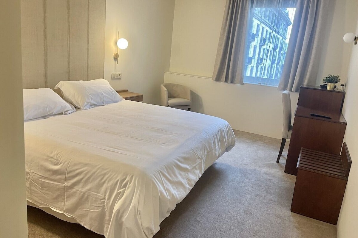 A hotel room with a large bed, a desk, a chair, and a window with curtains.