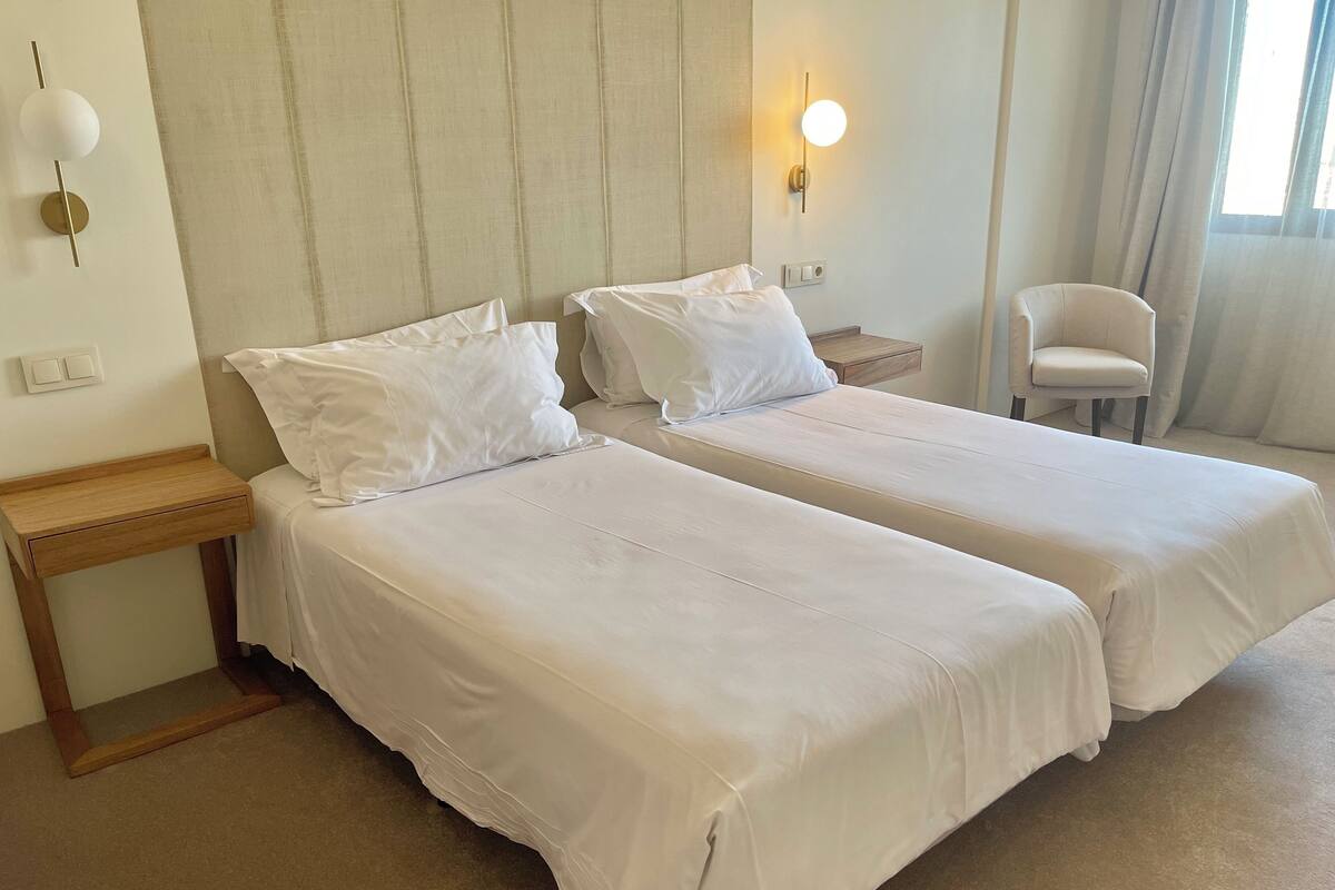 A hotel room with two beds, a wooden nightstand, a chair, and a wall-mounted light fixture.