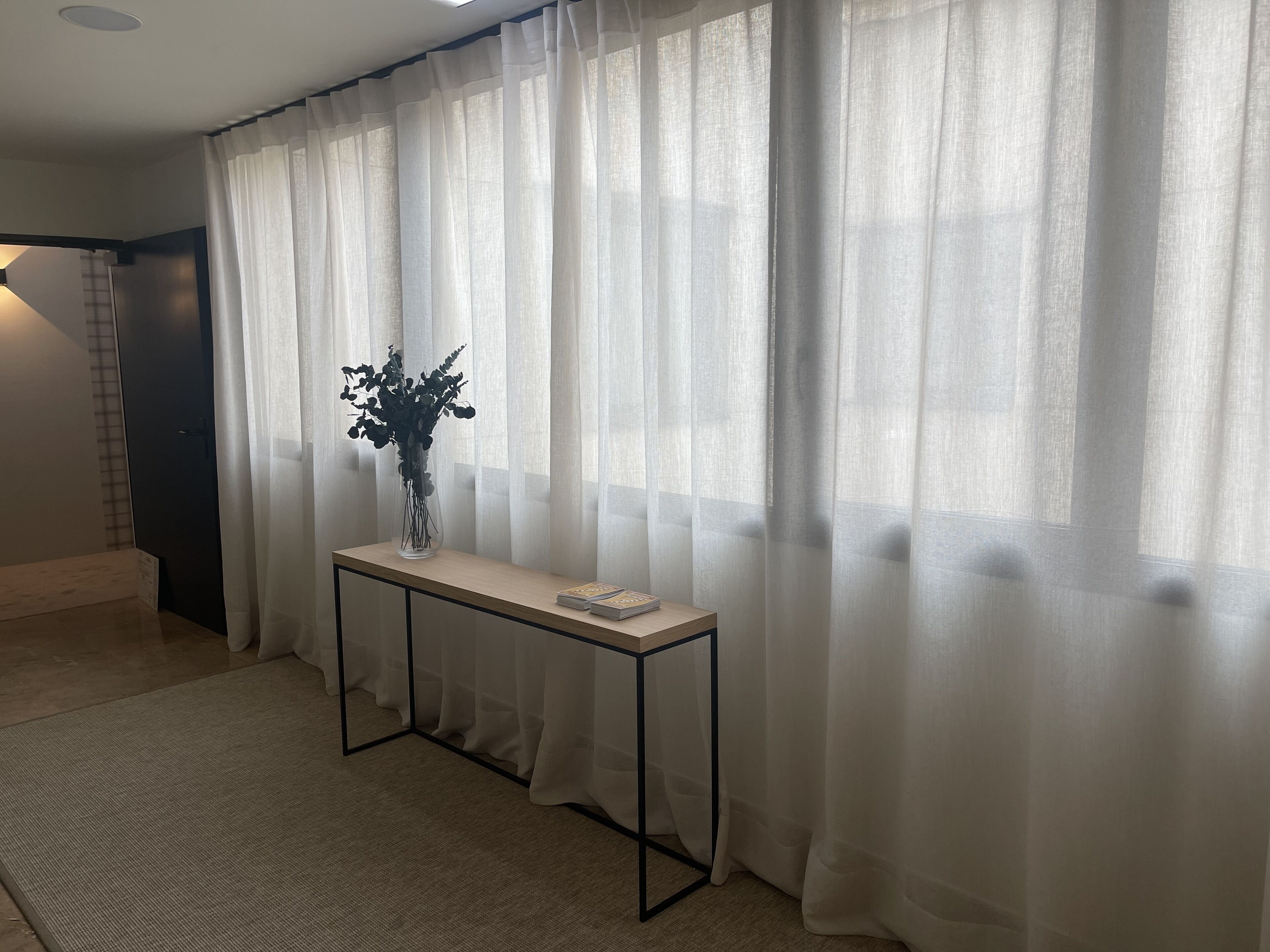 A room with a large window covered by sheer curtains, a small table with a vase of flowers, and a carpeted floor.