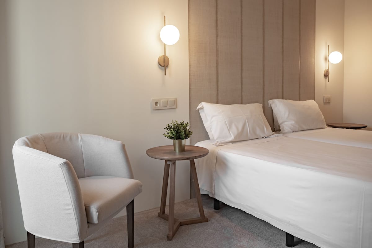 A hotel room with a bed, a chair, a small table with a plant, and wall-mounted lights.