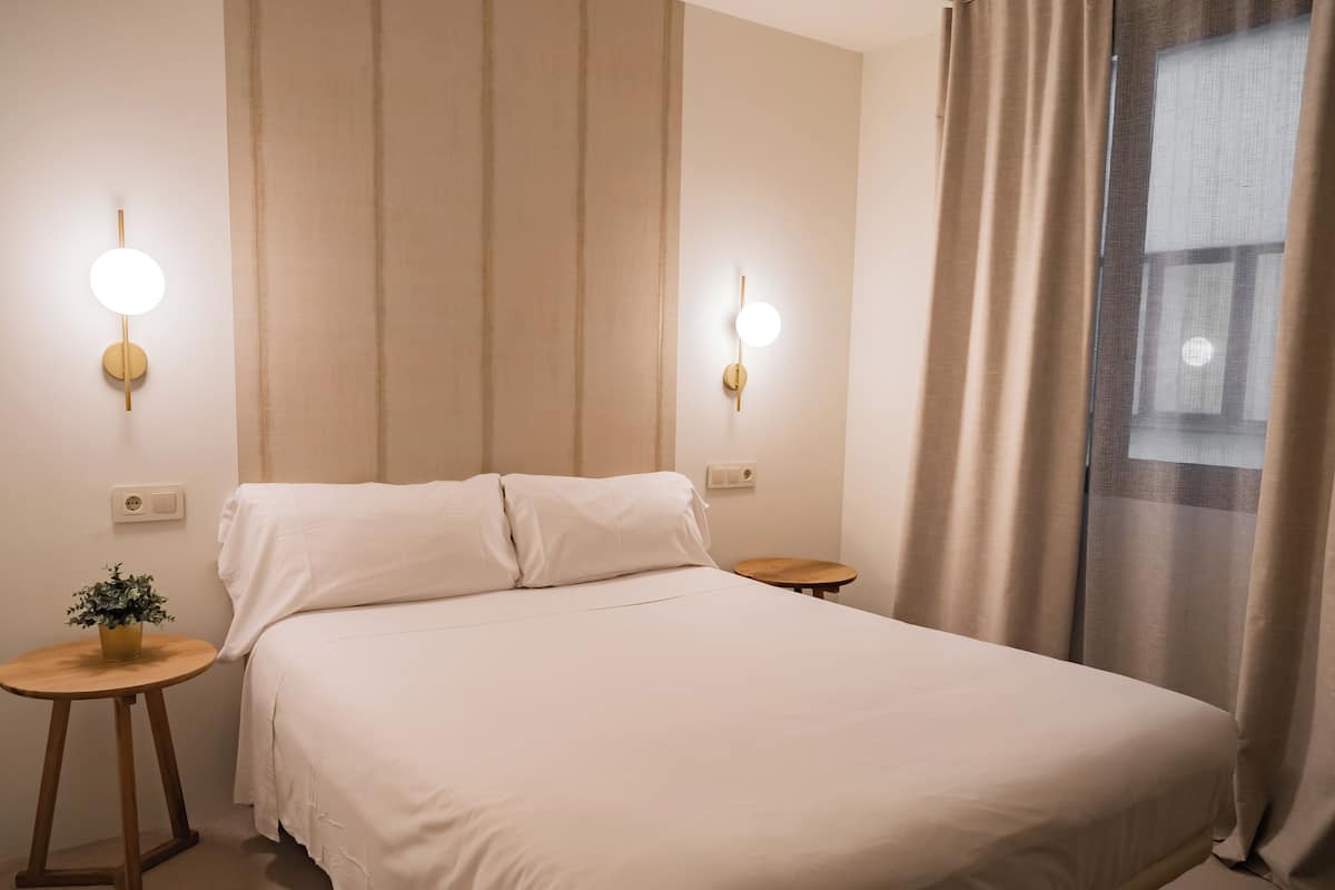 A neatly made bed with white linens, two bedside tables with lamps, a small potted plant, and a window with curtains.