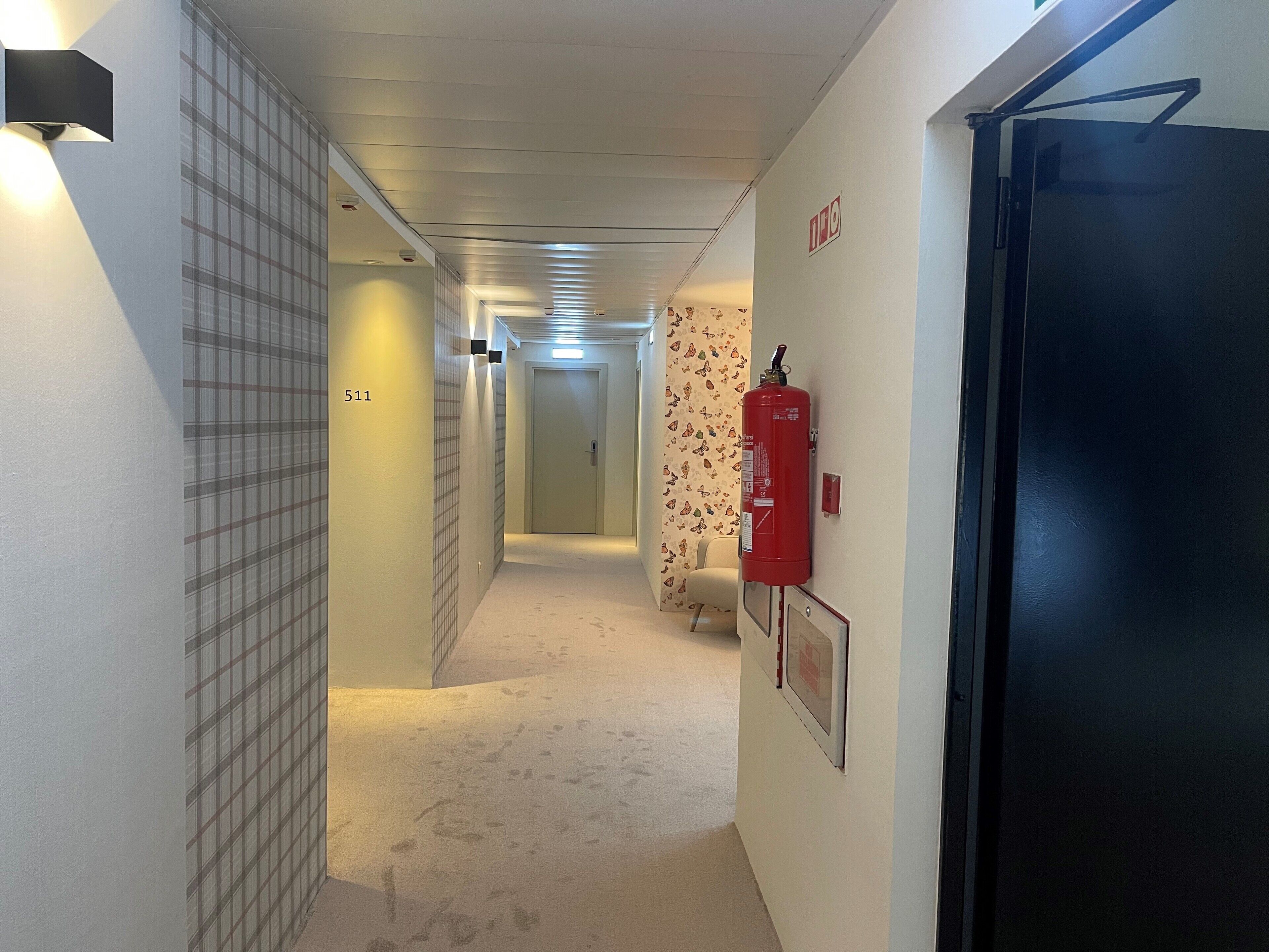 A hallway with a fire extinguisher mounted on the wall, a door with a number, and a patterned wallpaper.