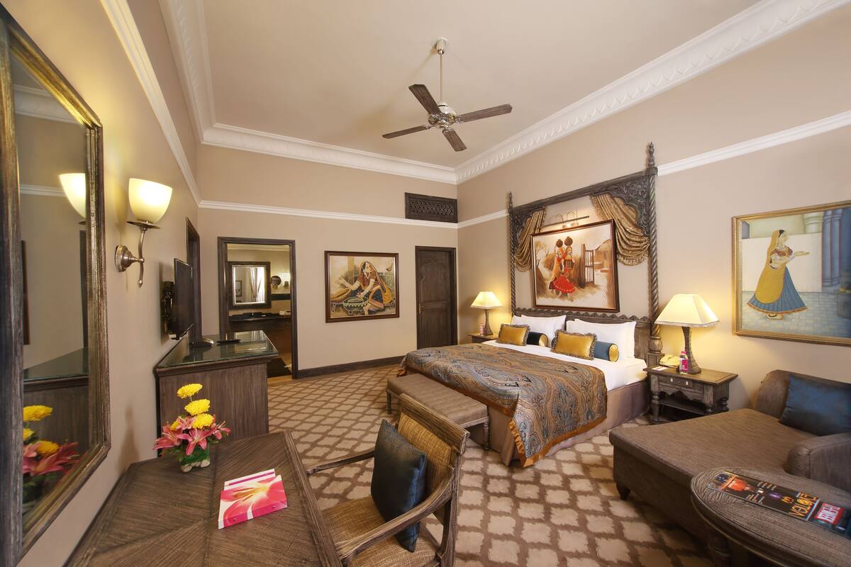 A hotel room with a bed, two bedside tables, a desk with a chair, a sofa, a ceiling fan, and a large mirror.