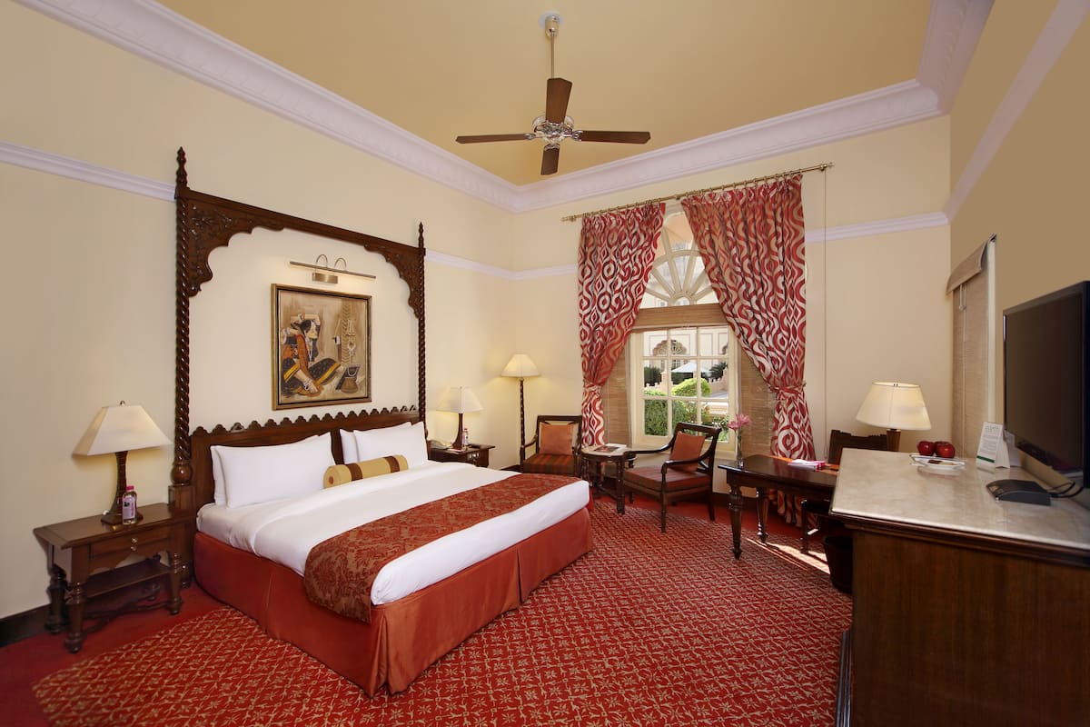 A hotel room with a large bed, two chairs, a desk, a television, and a ceiling fan.