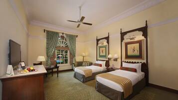 A hotel room with two beds, a desk with a fruit bowl, a desk lamp, a chair, a window with curtains, a ceiling fan, and framed artwork on the walls.