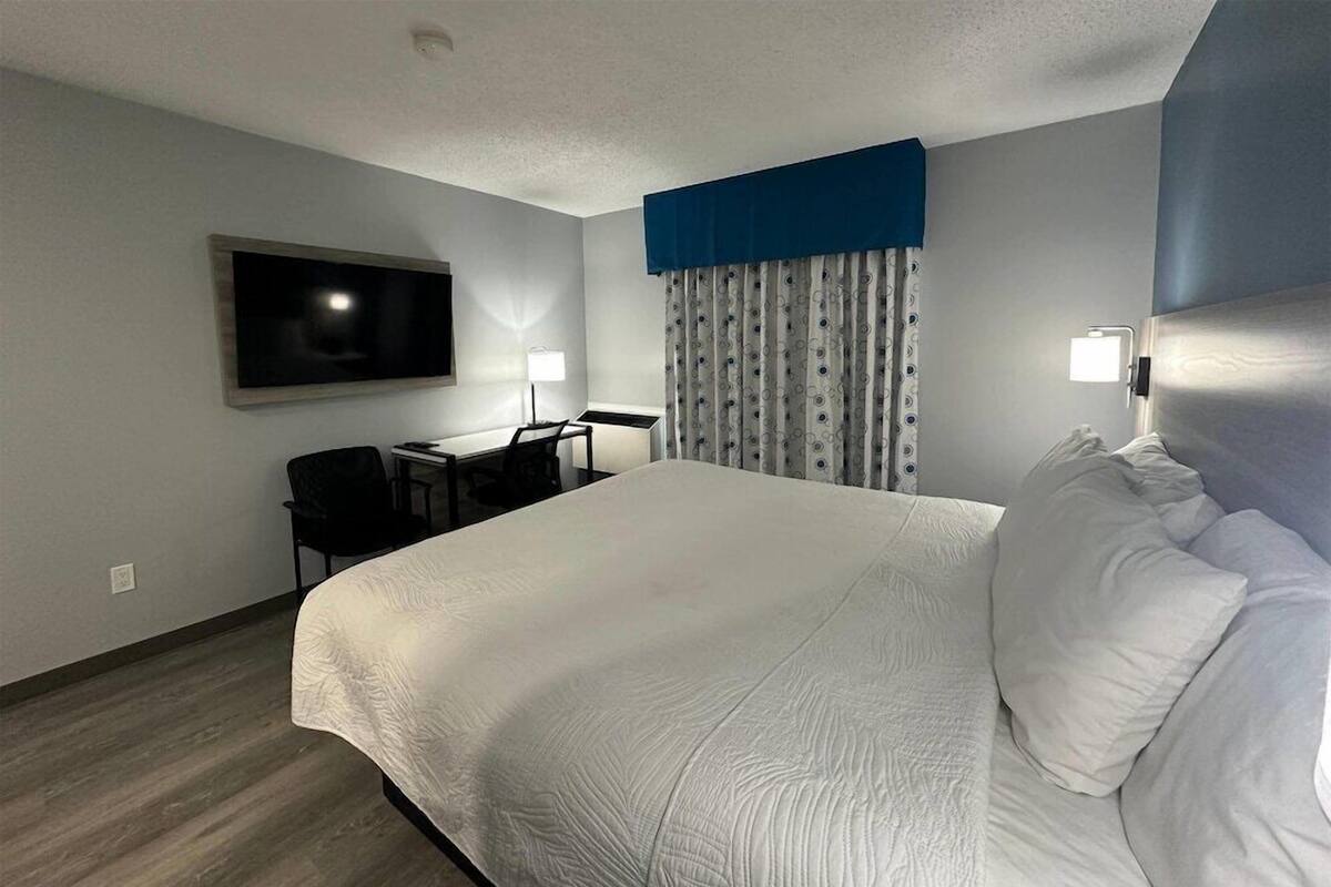Habitació d'hotel amb un llit gran, un escriptori amb una cadira, un televisor fixat a la paret i una finestra amb cortines.