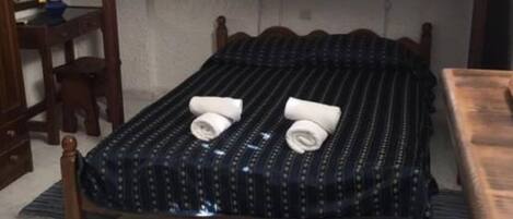 Ein Einzelbett mit einer dunkel gemusterten Tagesdecke und zwei gerollten Handtüchern darauf. Eine Holzstuhls und ein kleiner Tisch befinden sich links daneben. Ein dekorativer Teppich mit einem Drachenmuster liegt auf dem Boden.