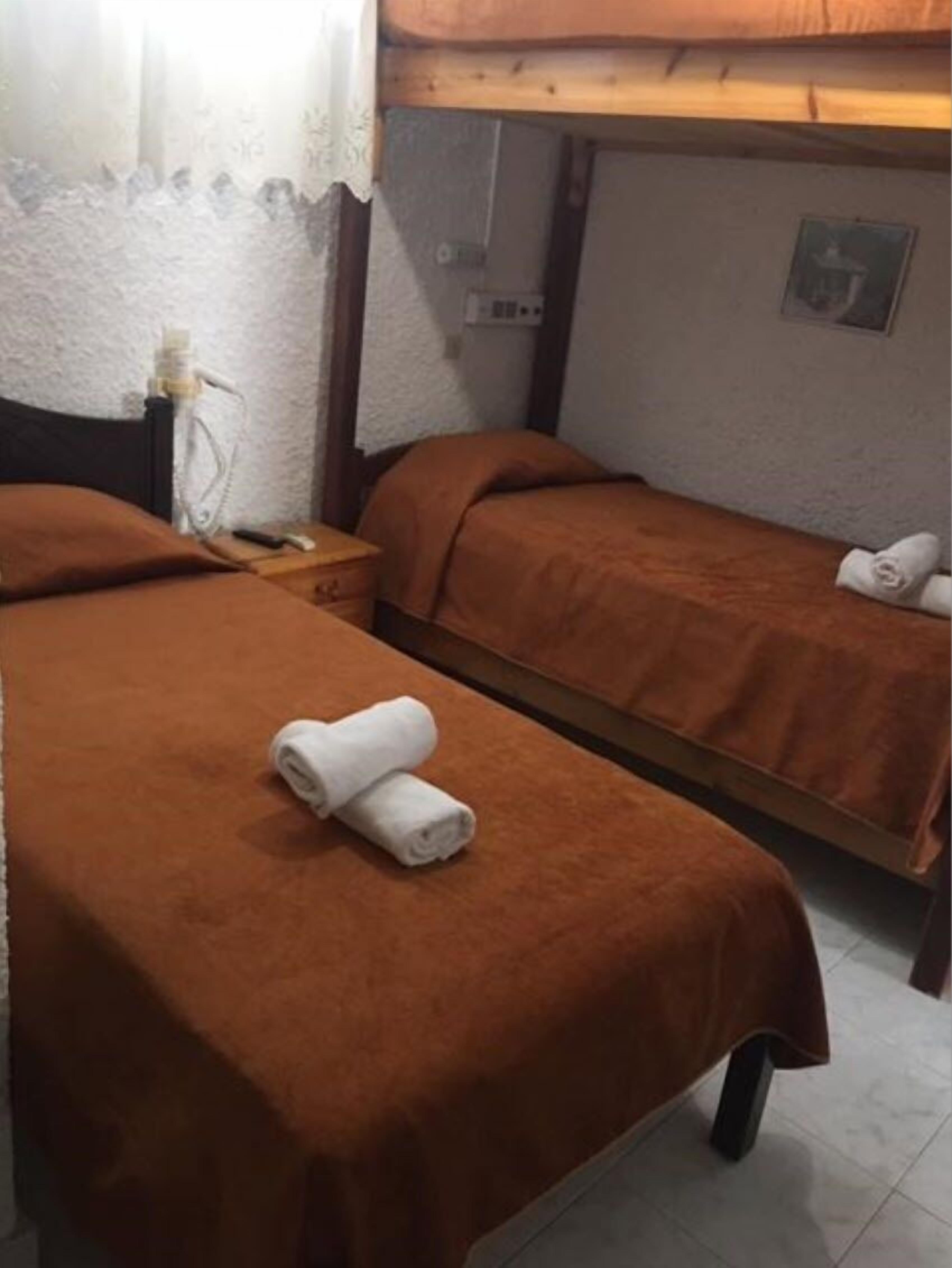 A small, cozy room with two single beds, each with a brown blanket and a rolled towel. There is a wooden headboard, a small table with a lamp, and a wall-mounted shelf with a picture frame.