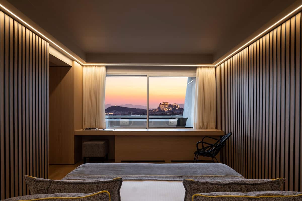 A modern hotel room with a large bed, a desk, and a view of a cityscape at sunset.