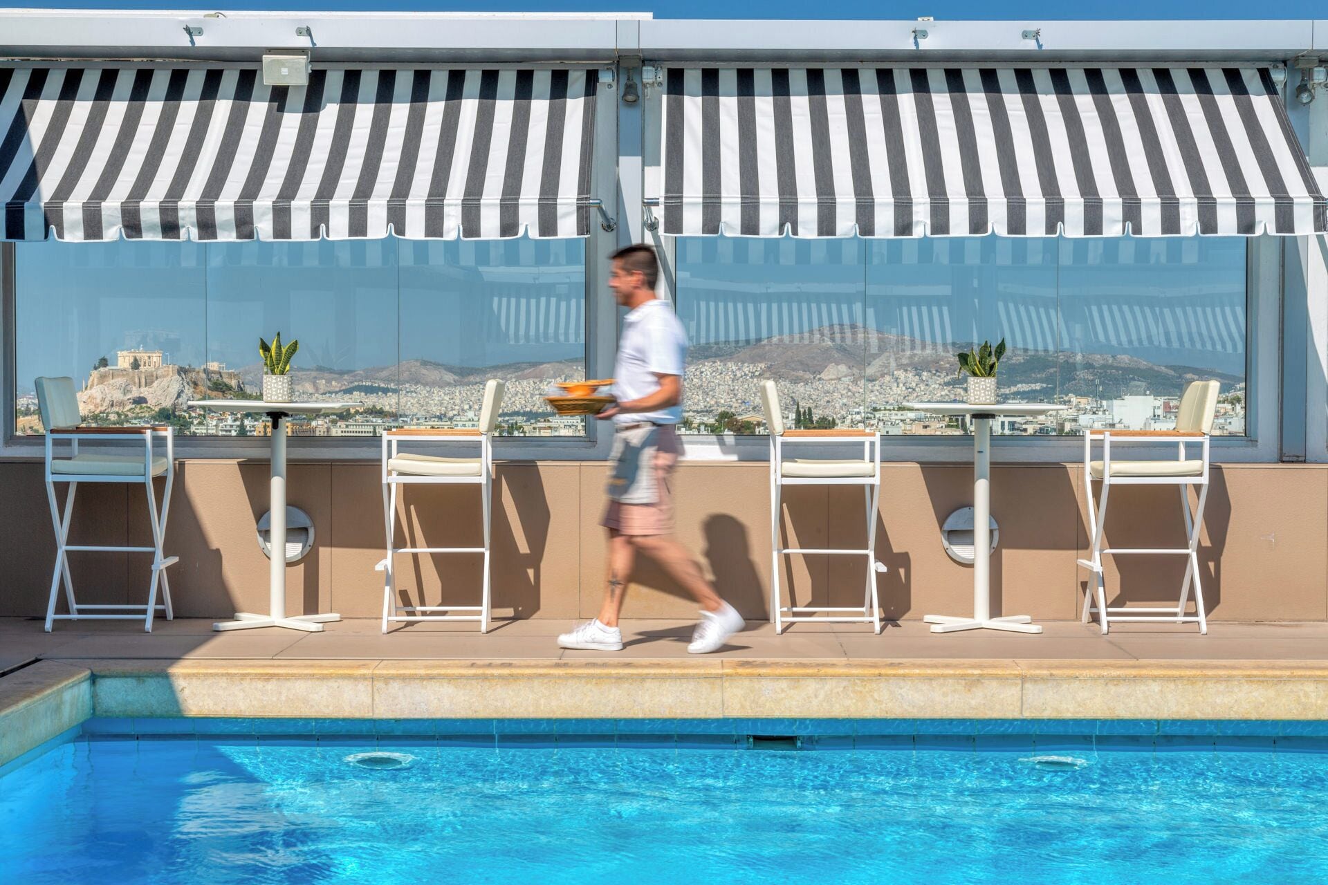 A poolside area with a view of a cityscape, including a historic building. There are white chairs and tables, and a person walking by.