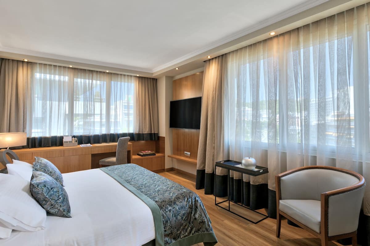 A hotel room with a bed, desk, chair, TV, and large windows with curtains.