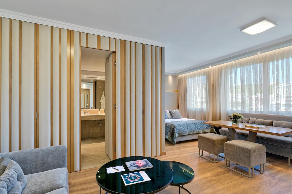 A modern hotel room with a bed, sofa, ottomans, a glass table, and a sliding door.