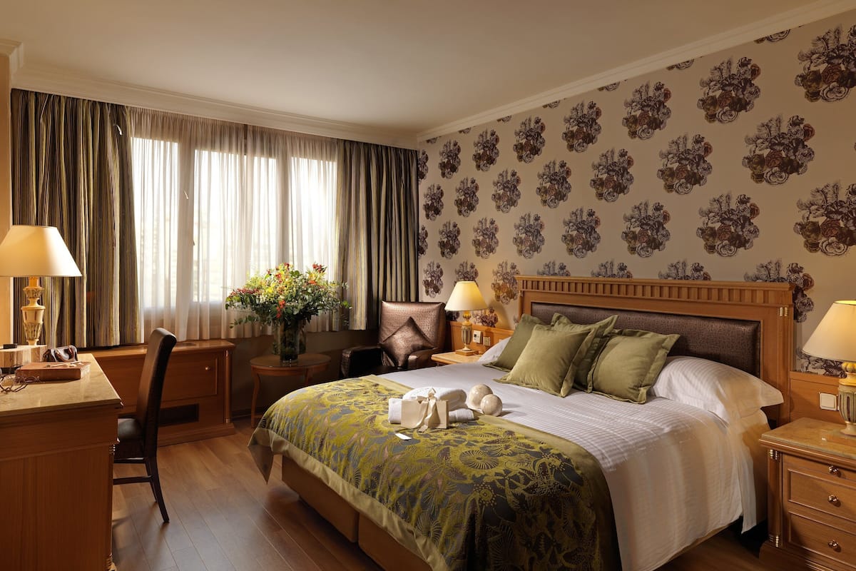 A hotel room with a bed, two bedside tables with lamps, a desk with a chair, a large window with curtains, and a floral wallpaper pattern.
