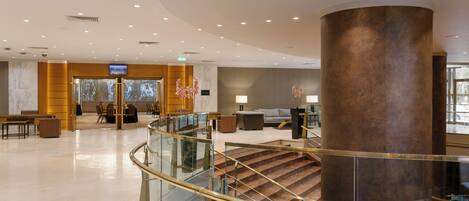A spacious lobby with a marble floor, a large central column, and a staircase with glass railings.