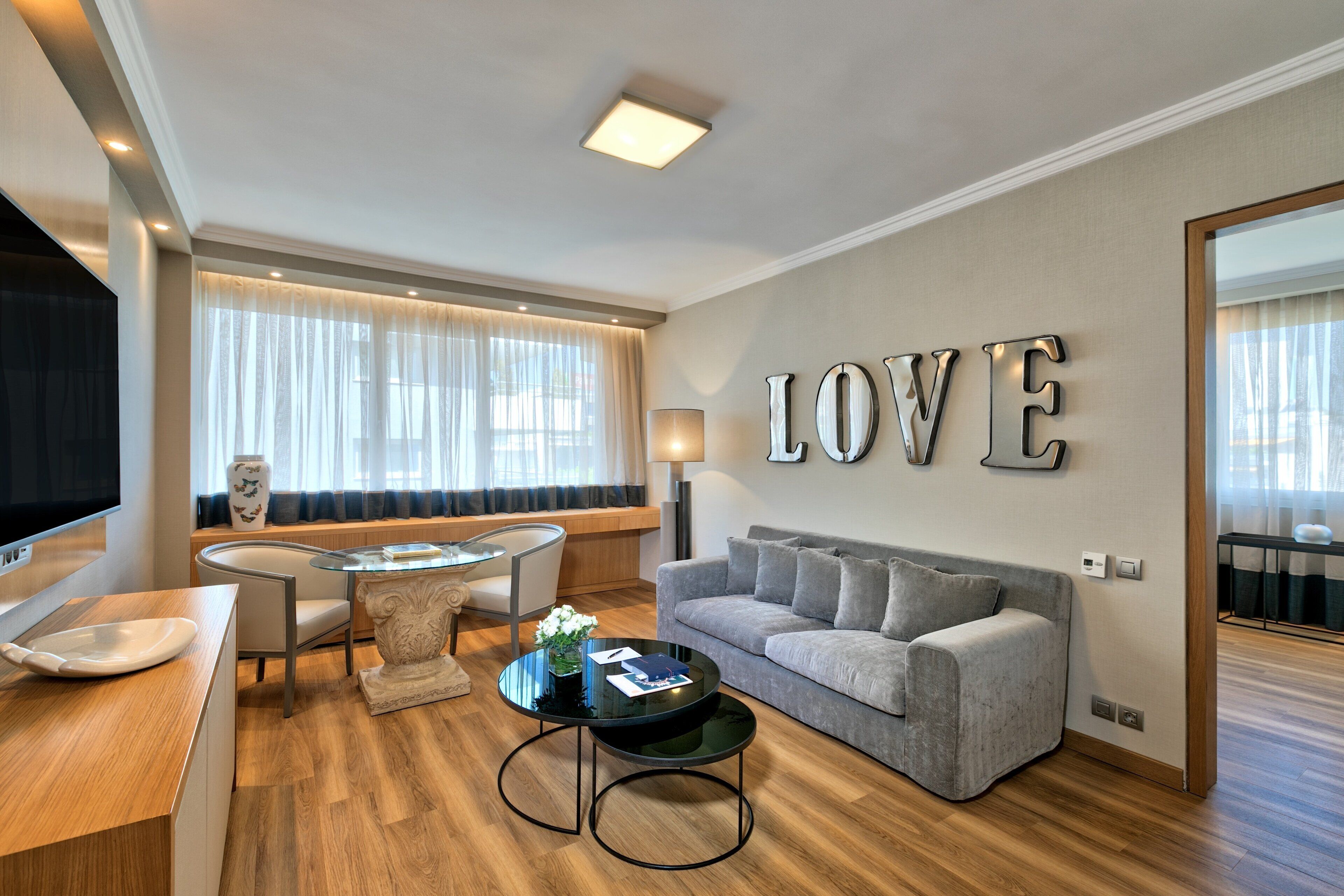 Een moderne woonkamer met een bank, een glazen salontafel en een televisie. Op de muur staat een groot 'LOVE'-bord en op een bijzettafeltje staat een lamp.
