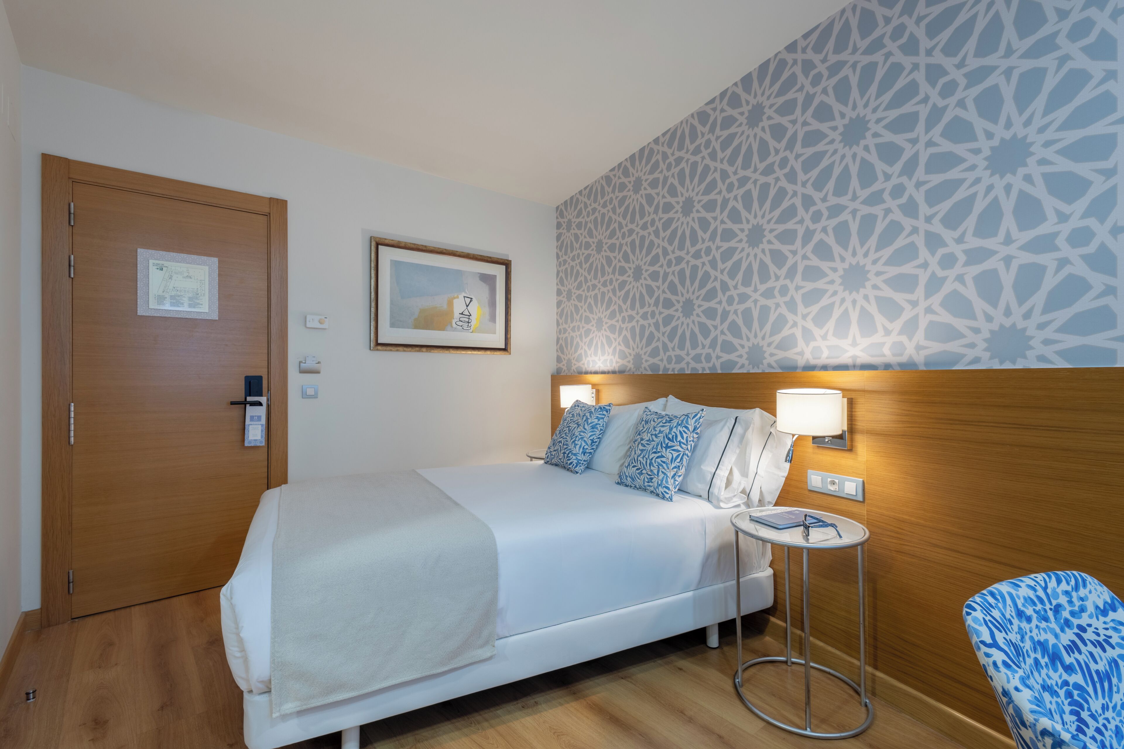 Habitación de hotel con cama, mesita de noche, silla y pared con estampado.
