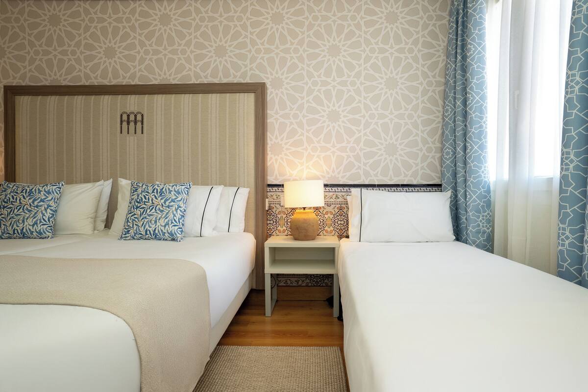 Habitación de hotel con dos camas, una mesita de noche con lámpara, papel tapiz estampado y vistas al exterior a través de cortinas.