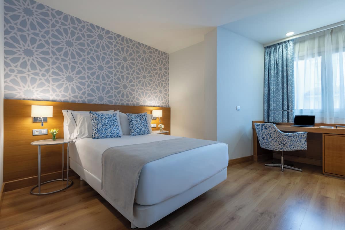Una habitación de hotel moderna con una cama grande, mesitas de noche, un escritorio con una silla y una pared con diseño.