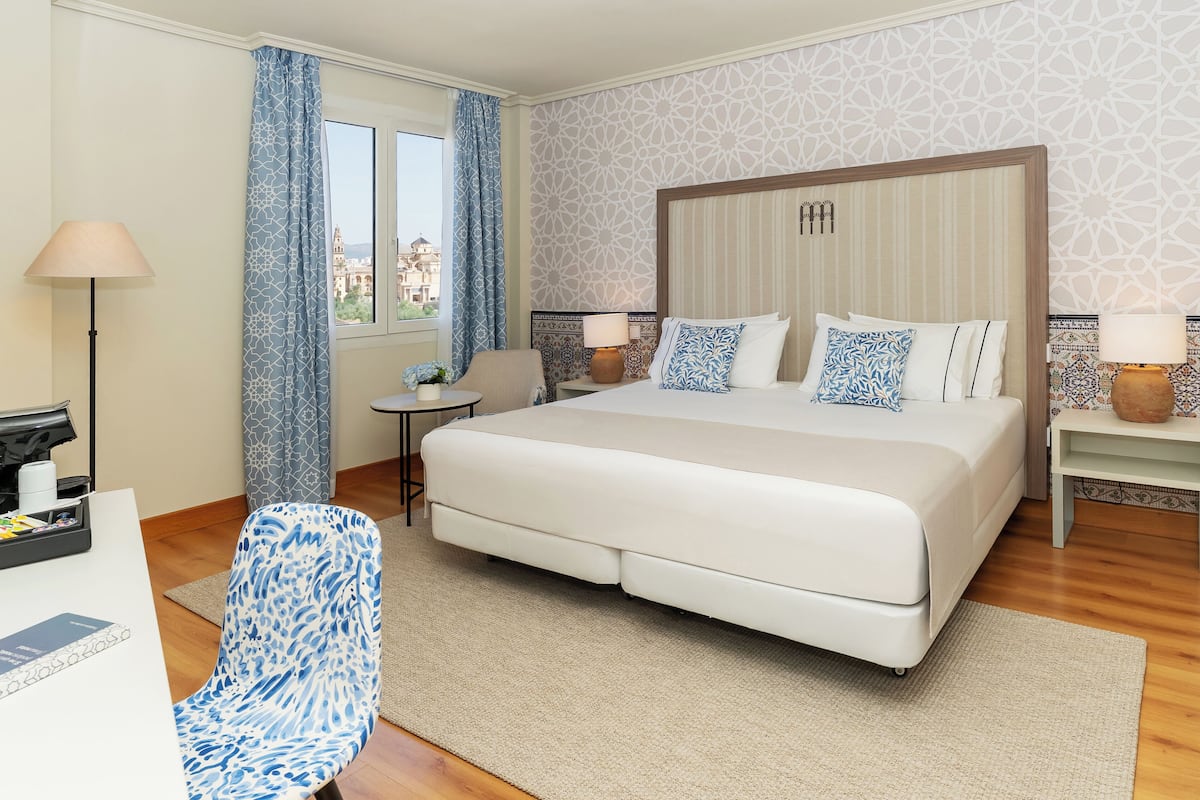 Habitación de hotel con una cama grande, un escritorio con lámpara, una silla y vistas a un paisaje urbano.