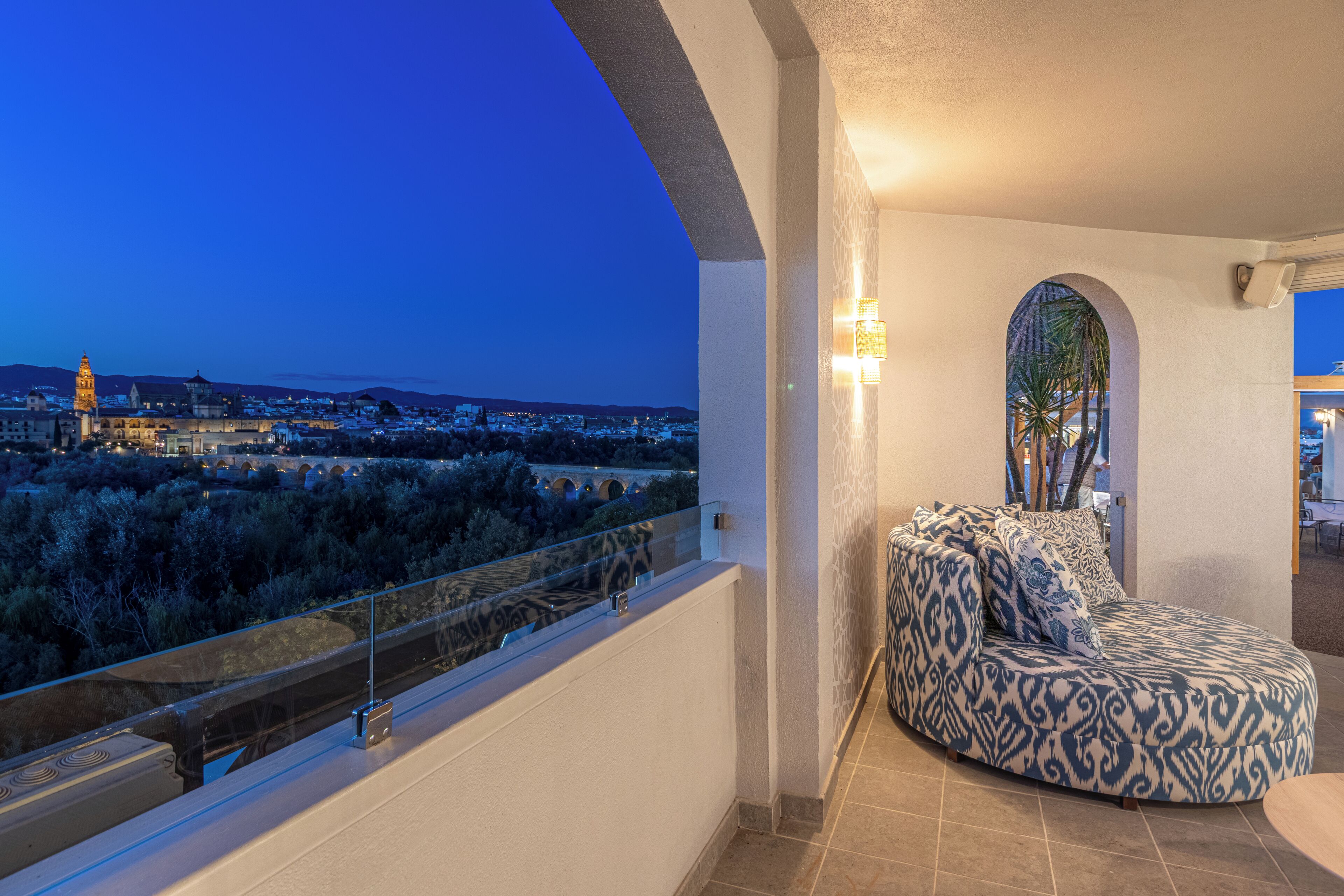 A balcony with a curved sofa, a glass railing, and a view of a cityscape at dusk.