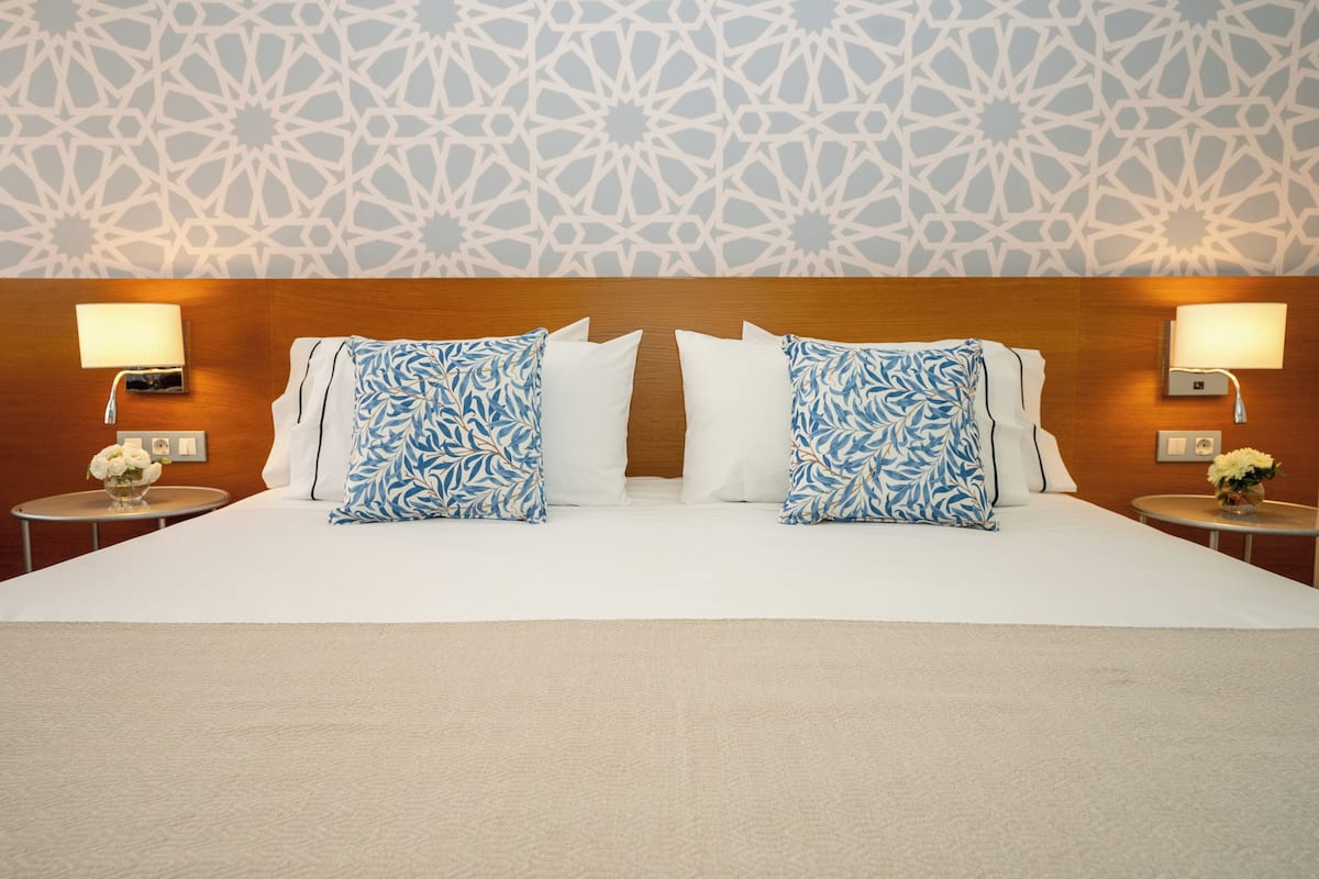 Habitación de hotel con una cama grande, dos almohadones estampados, un cabecero de madera y dos mesitas de noche con lámparas.