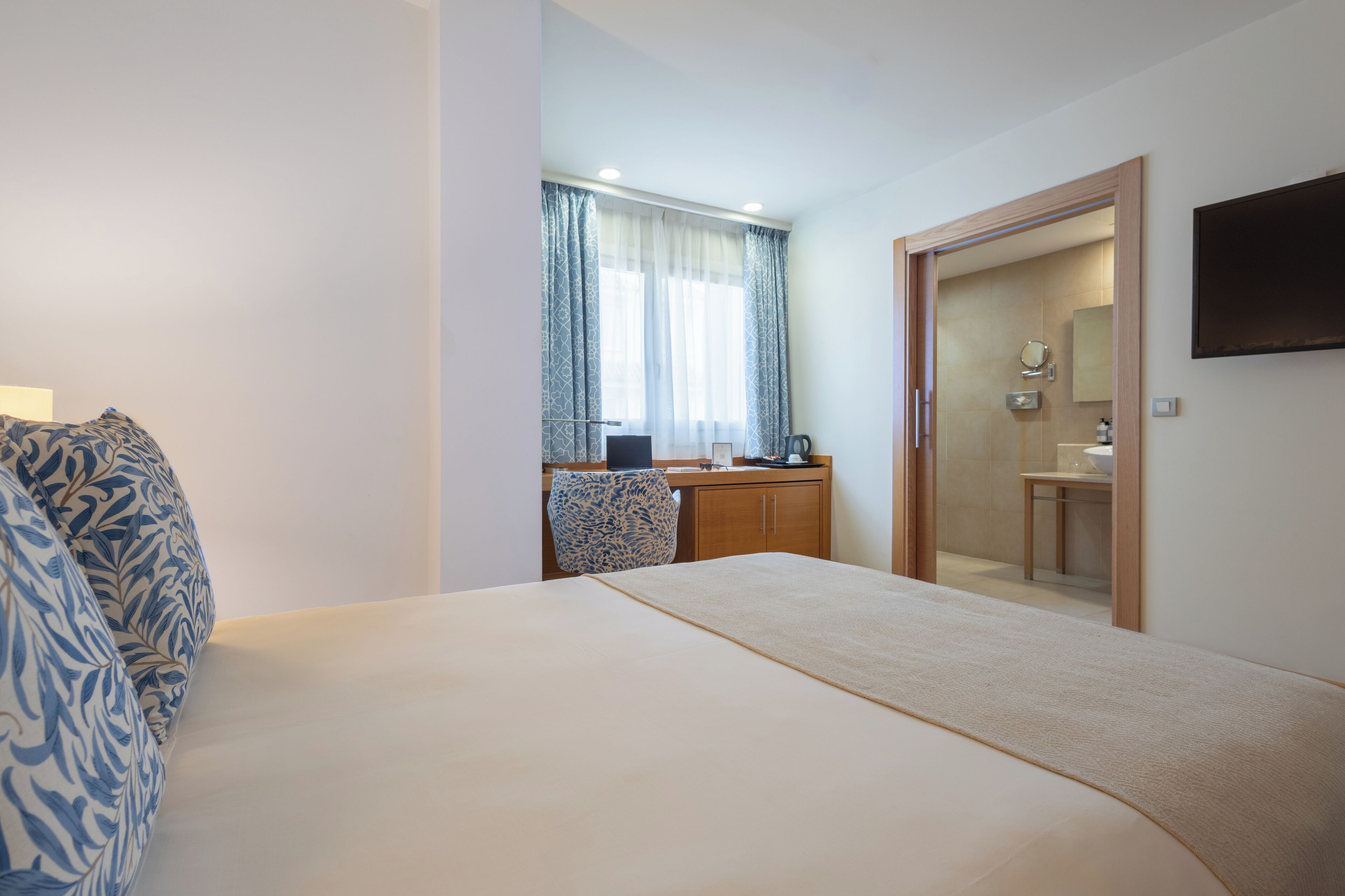 A hotel room with a large bed, a desk with a chair, a mirror, and a TV mounted on the wall.