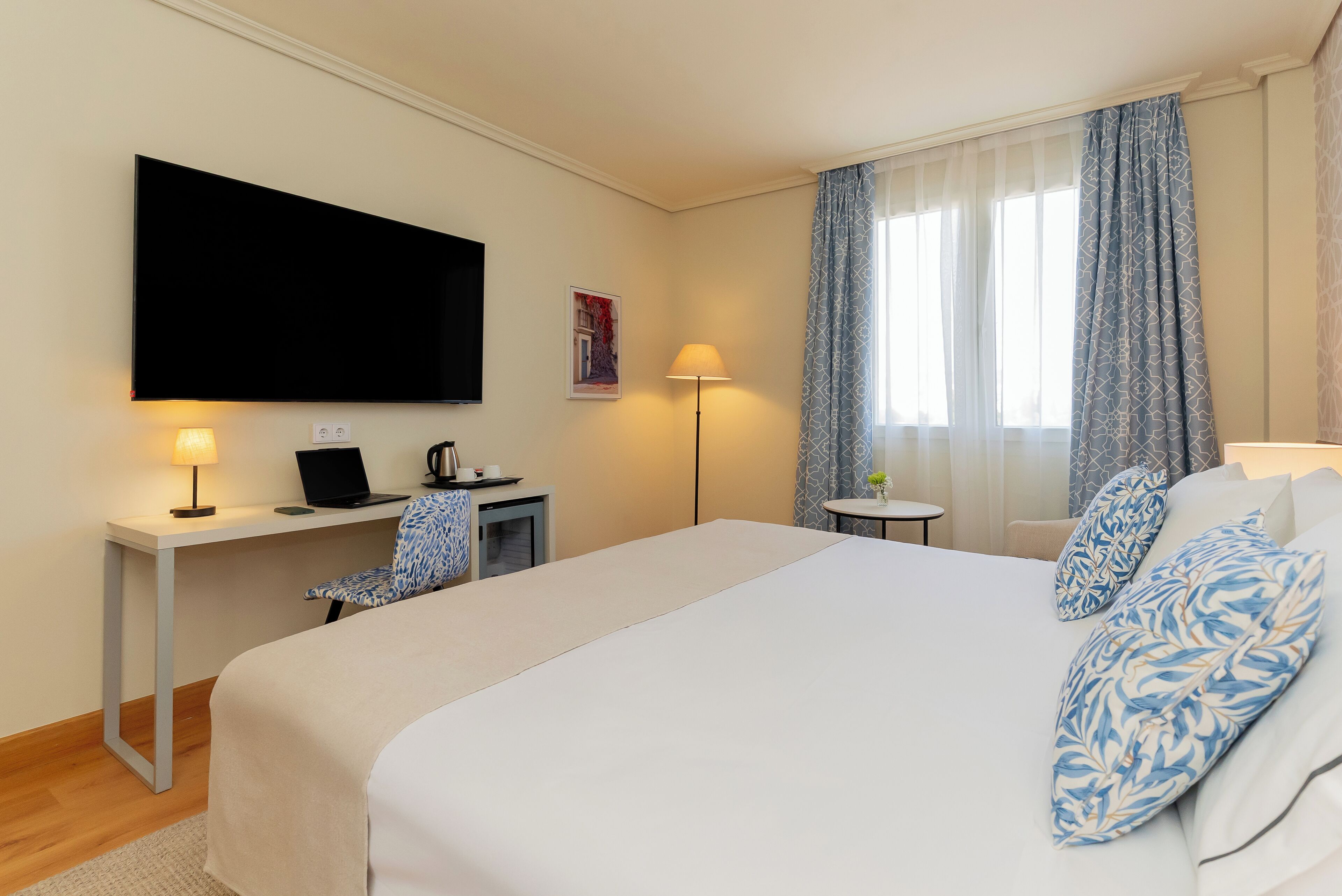 A hotel room with a large bed, a desk with a laptop, a flat-screen TV, a small table with a lamp, and a window with curtains.