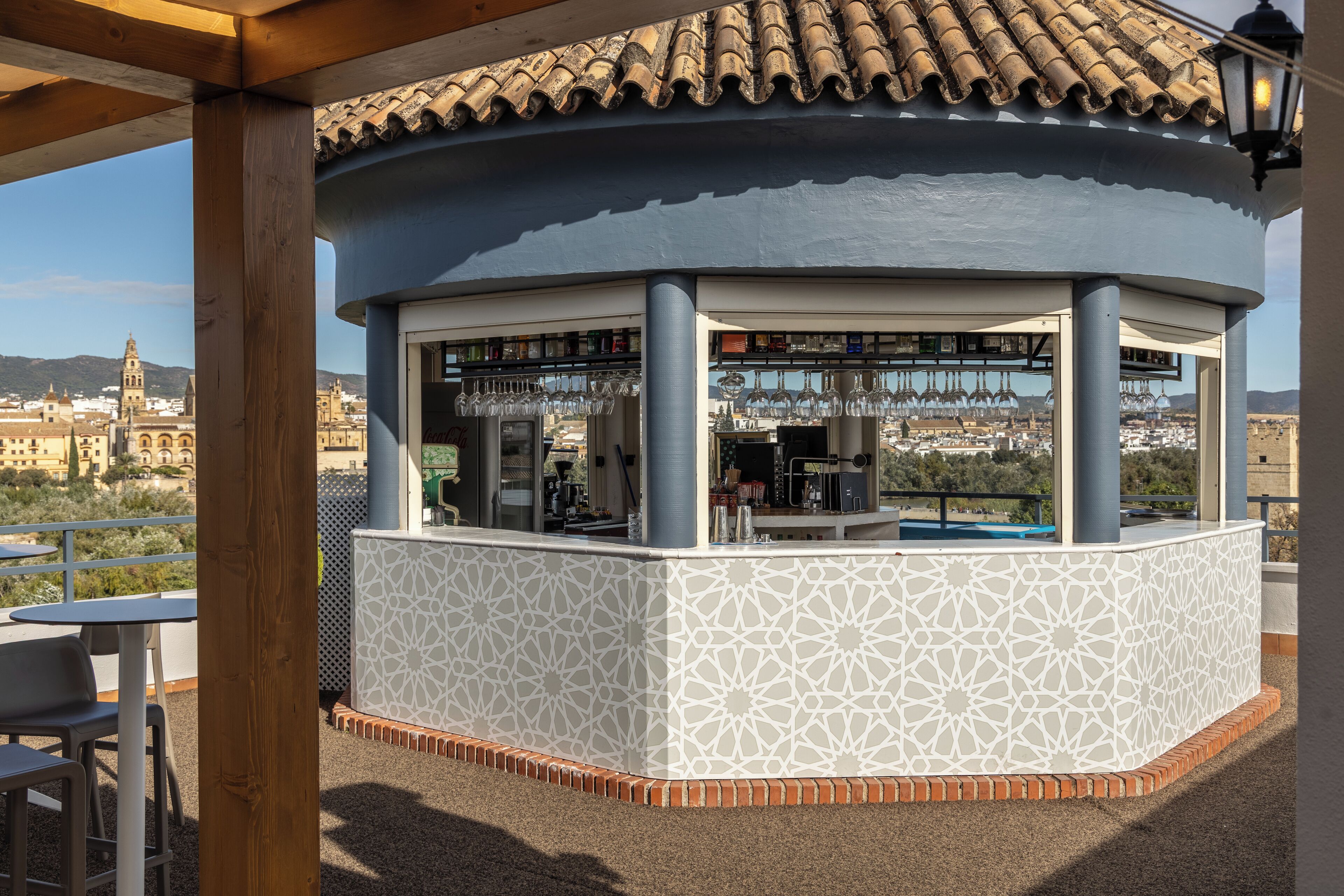 Un bar extérieur abrité, doté d’une façade blanche à motifs, d’un toit en tuiles et offrant une vue sur le paysage urbain en arrière-plan.