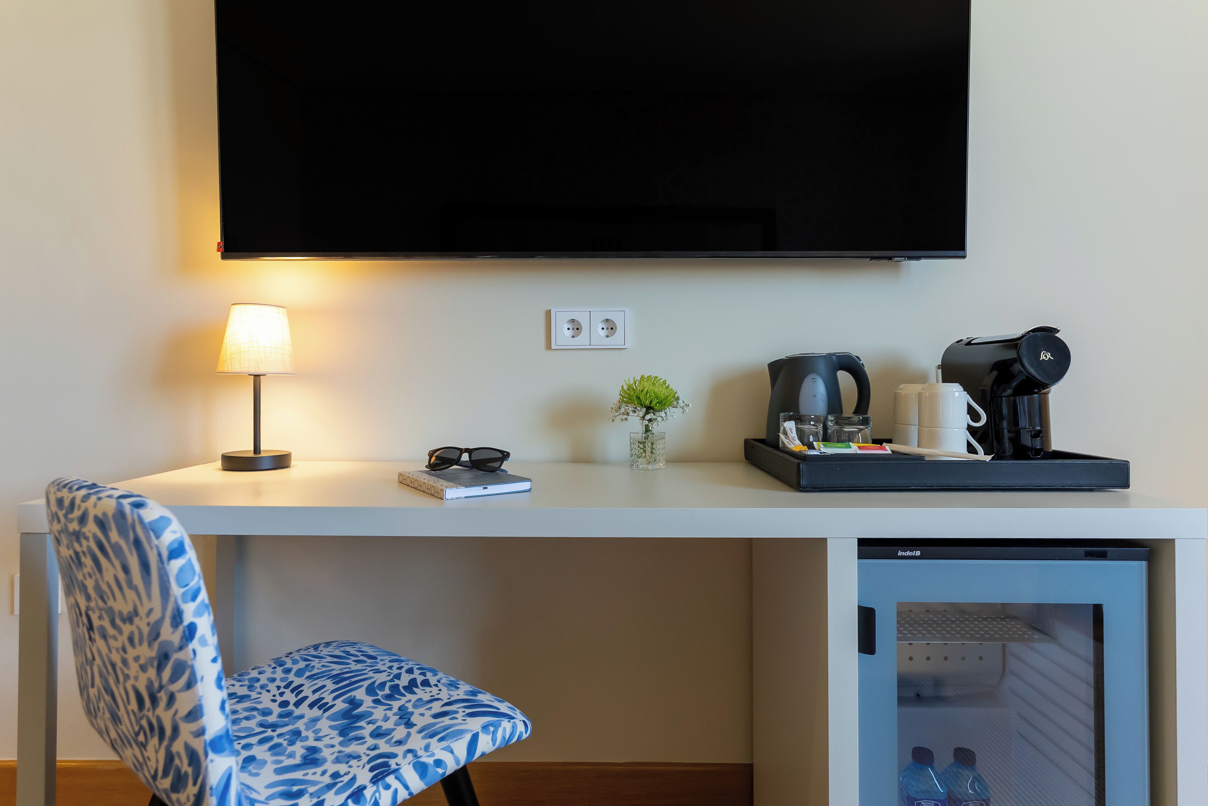 Ein Hotelzimmer mit Flachbildfernseher an der Wand, einem Schreibtisch mit Lampe, einer Kaffeemaschine, einer kleinen Pflanze und einem Kühlschrank.