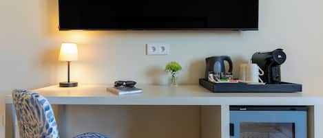 A hotel room with a flat-screen TV mounted on the wall, a desk with a lamp, a coffee maker, a small plant, and a refrigerator.