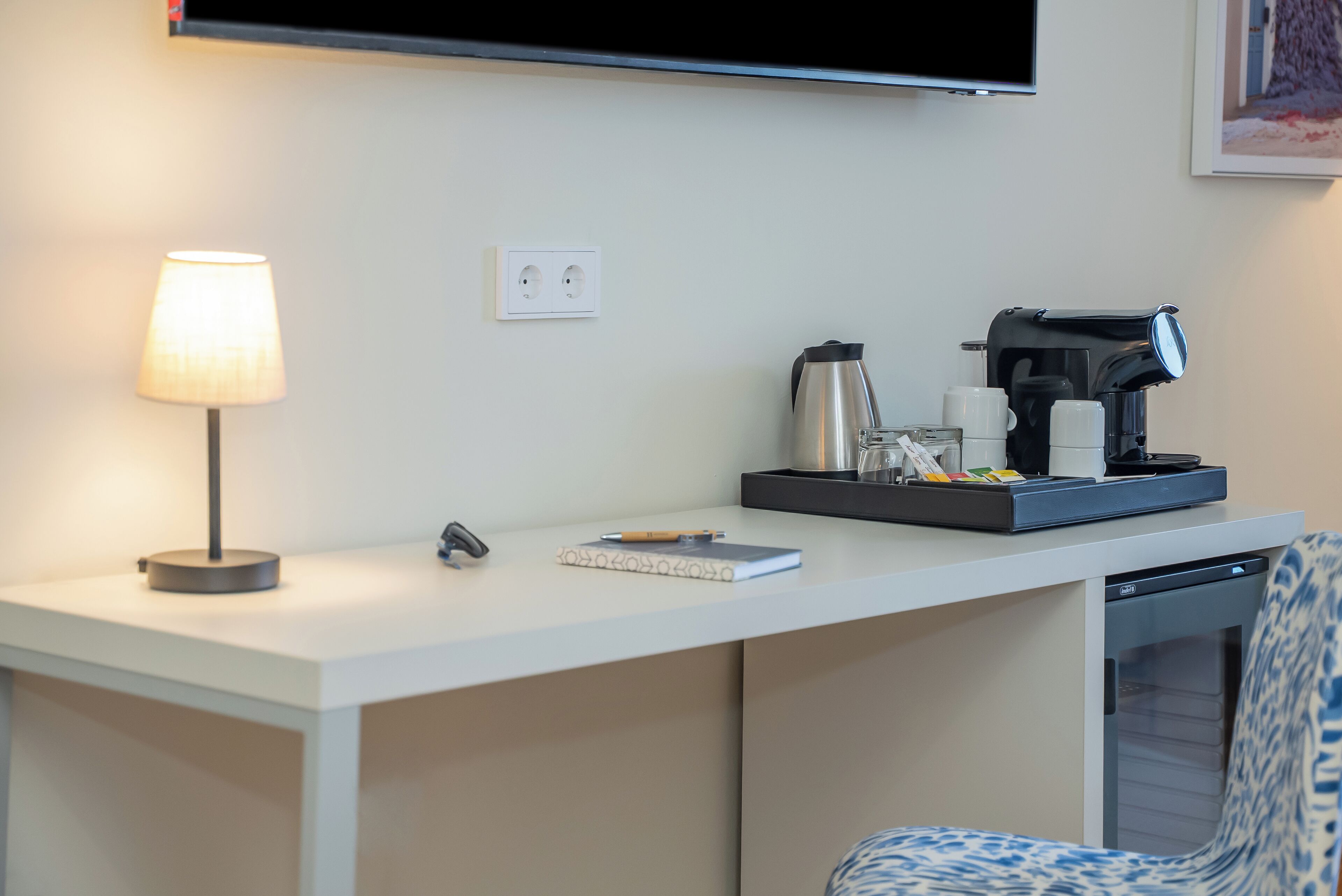 Una habitación de hotel con un escritorio blanco, una lámpara, cafetera, hervidor de agua y un cuaderno.