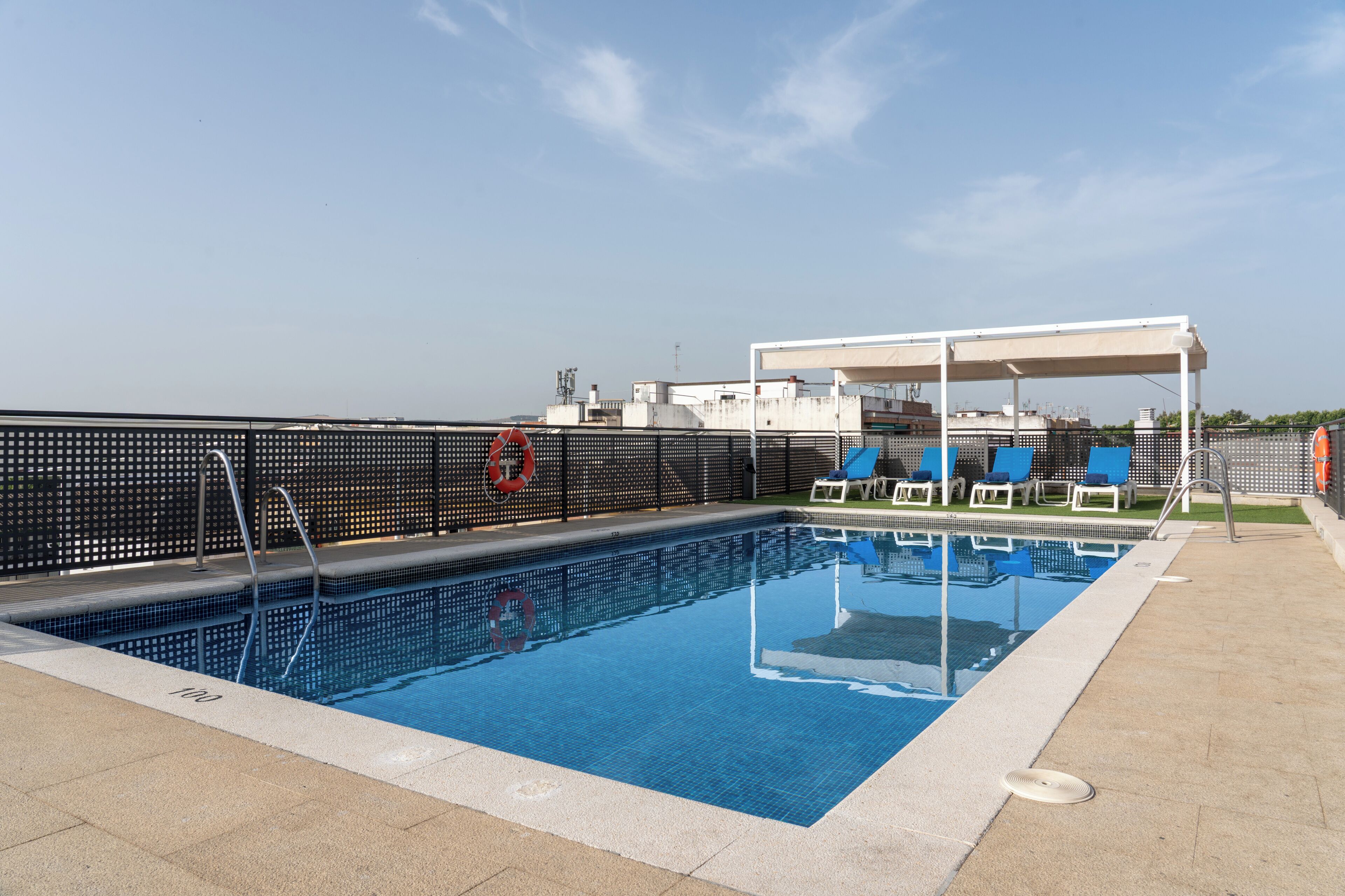 A rooftop swimming pool with a clear blue water surface, surrounded by a black mesh fence, and equipped with blue lounge chairs and a white canopy structure.
