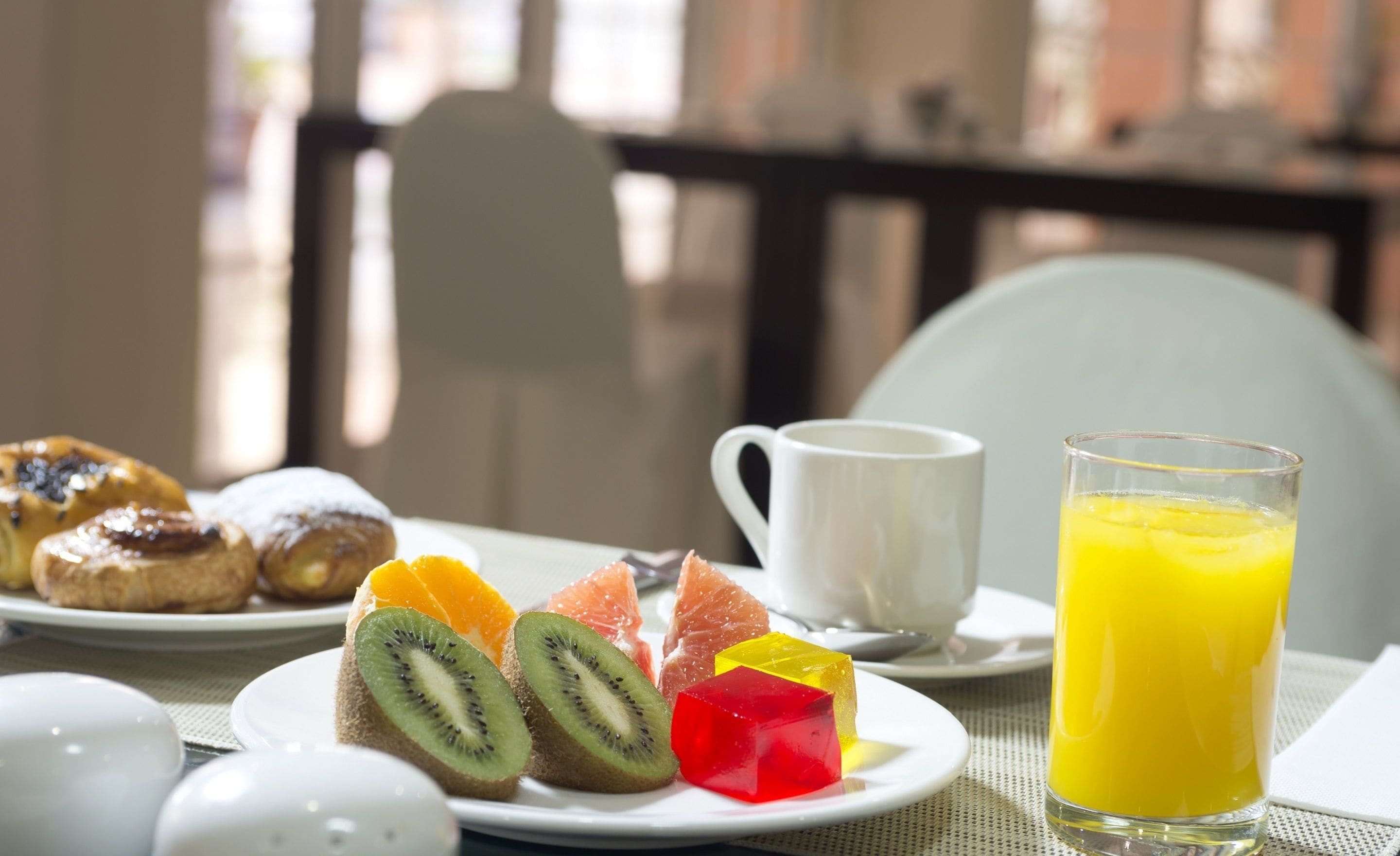 Un desayuno con pasteles, fruta, una taza y un vaso de zumo de naranja sobre una mesa.