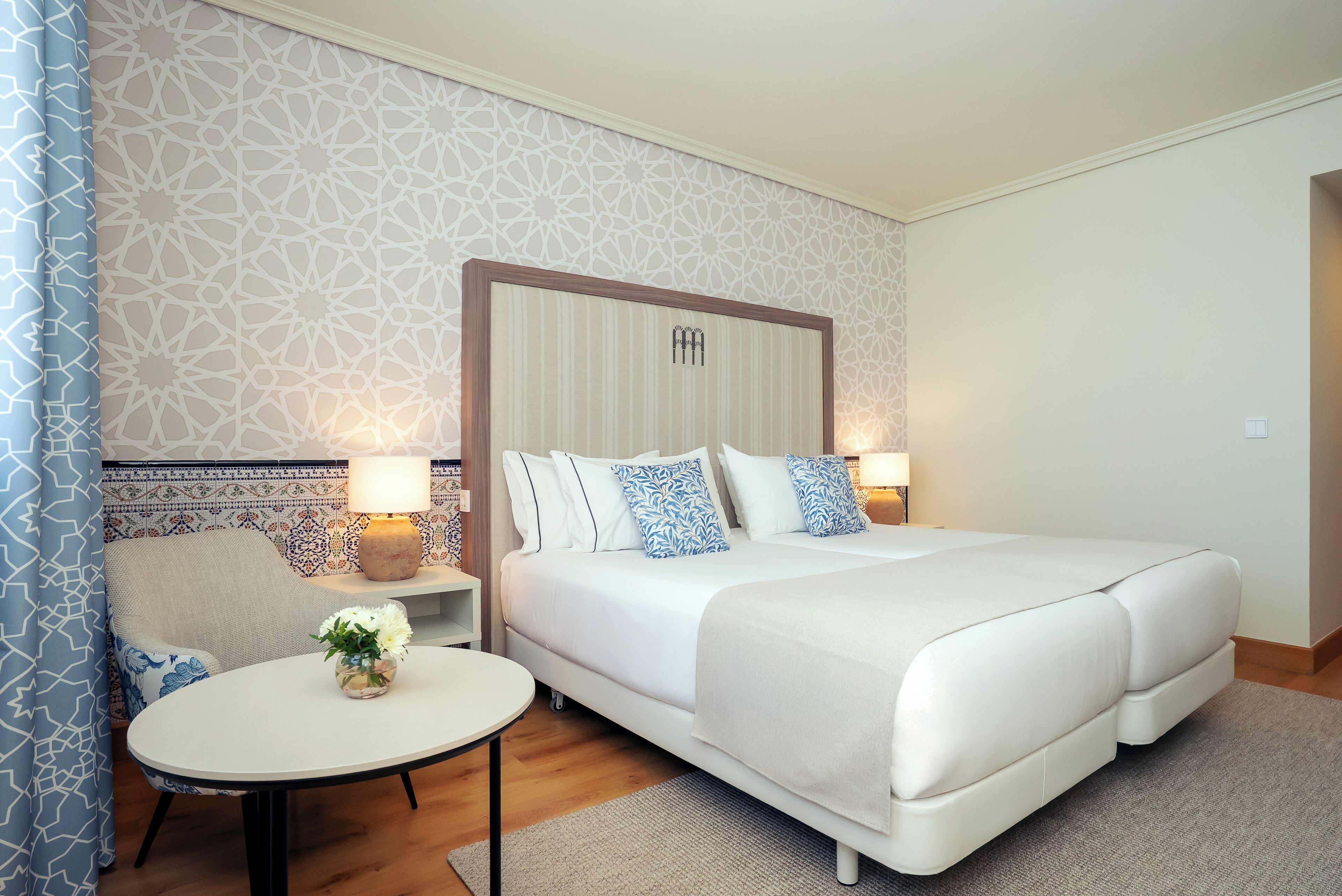 A hotel room with a large bed, a bedside table with a lamp, a small round table with a vase of flowers, and a patterned wall.