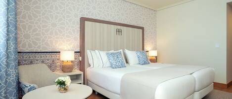 A hotel room with a large bed, a bedside table with a lamp, a small round table with a vase of flowers, and a patterned wall.