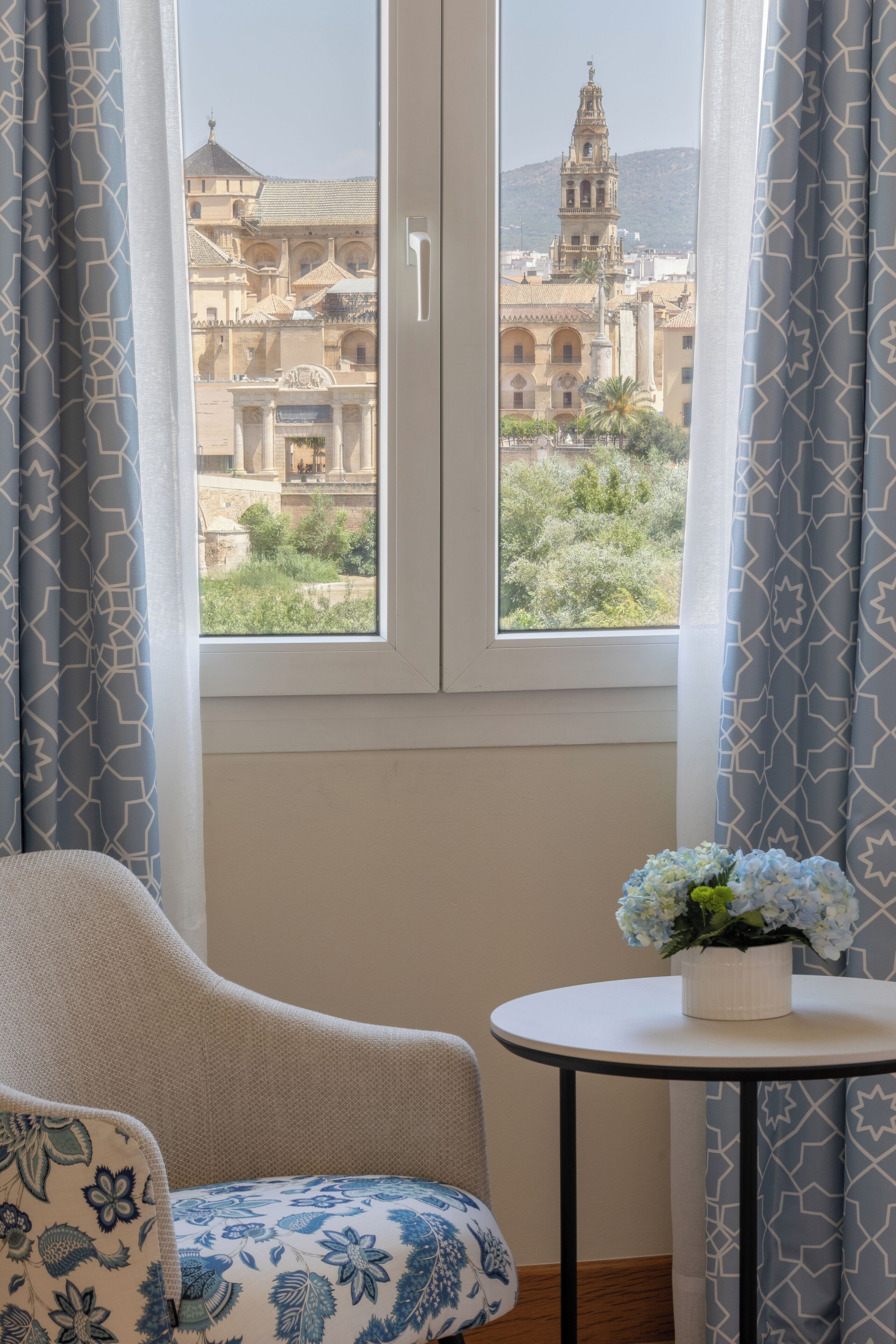 A room with a view of a historic building through a window, a chair, a small table with a potted plant, and patterned curtains.