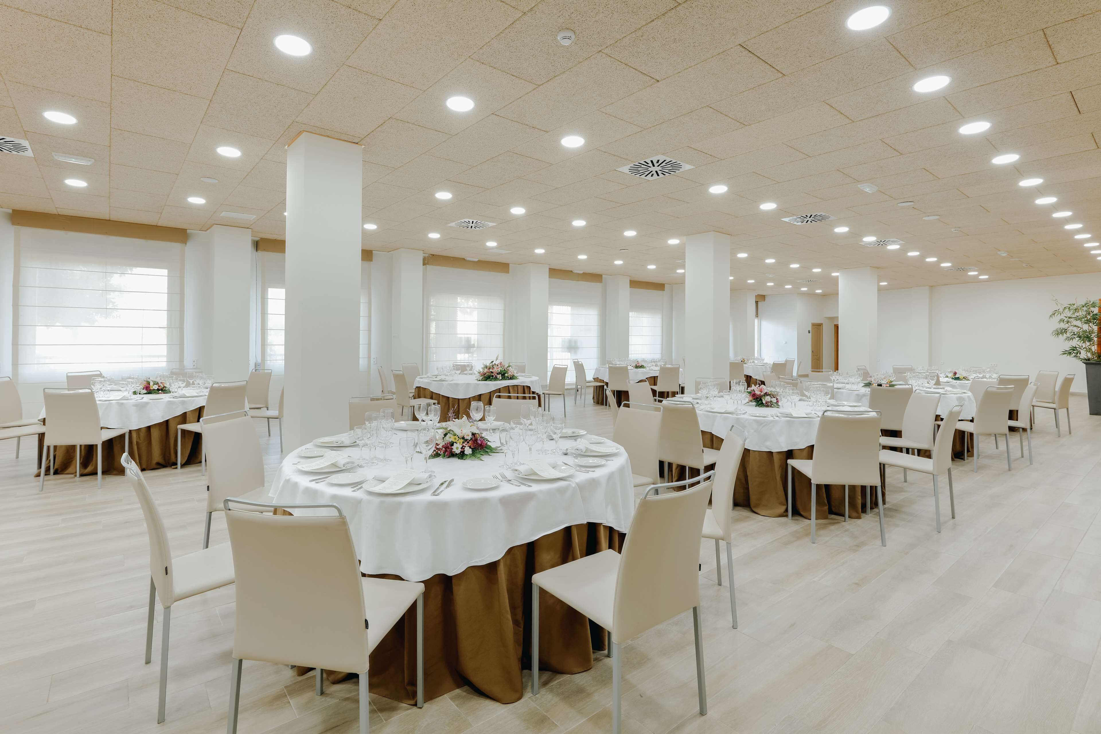 A spacious banquet hall with multiple round tables set for dining, featuring white tablecloths and gold drapes.