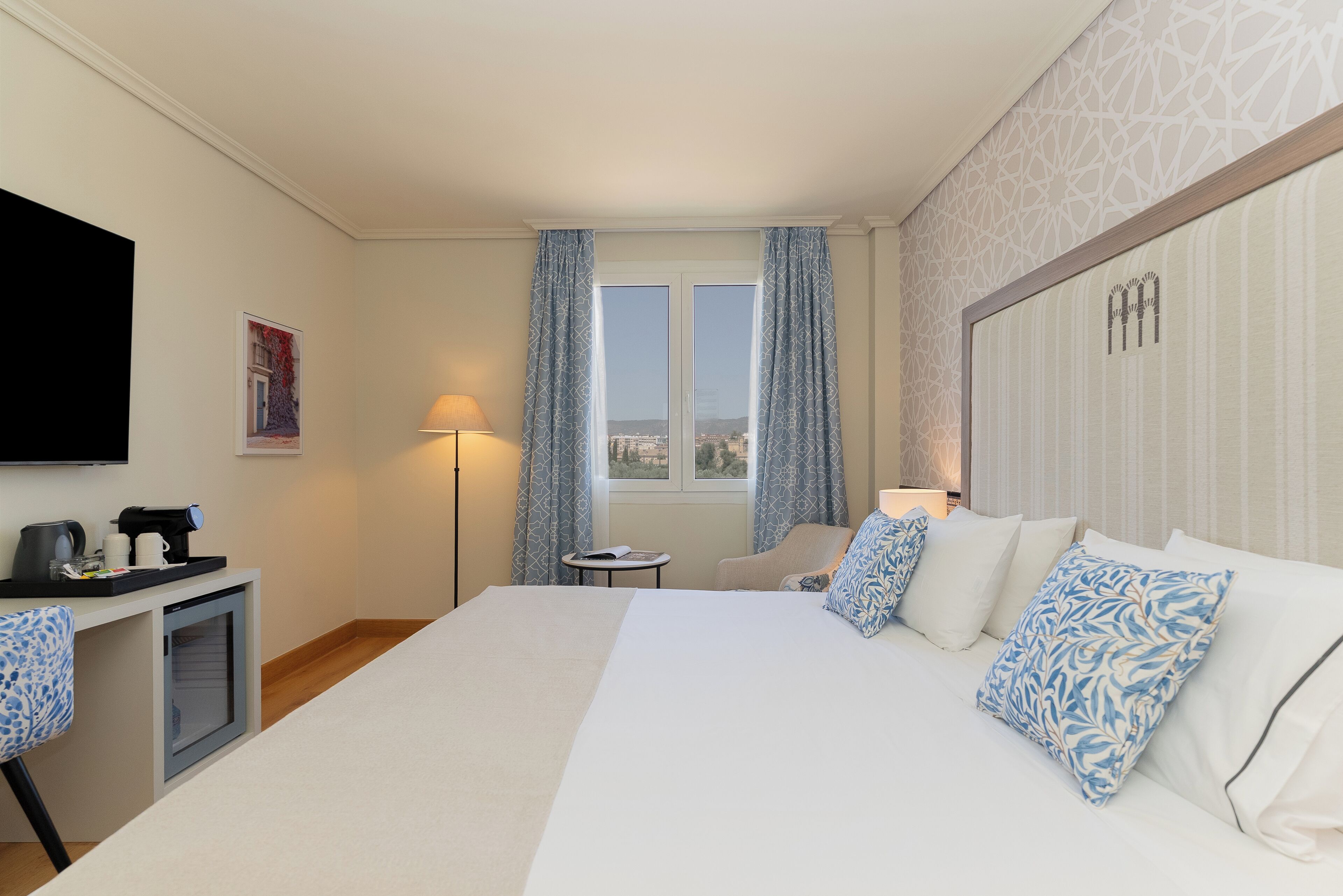 Une chambre d’hôtel avec un grand lit, un bureau avec une machine à café, une chaise, un téléviseur fixé au mur et une fenêtre avec des rideaux.