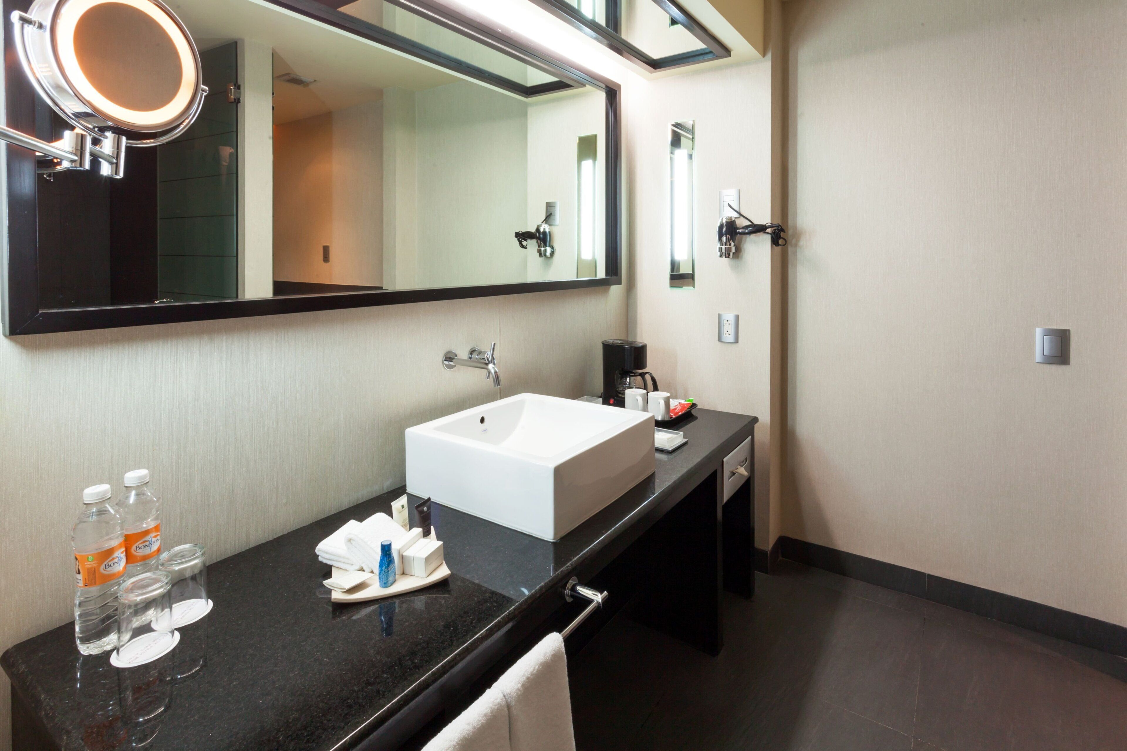 A hotel bathroom with a large mirror, a white sink, a black countertop, and toiletries.