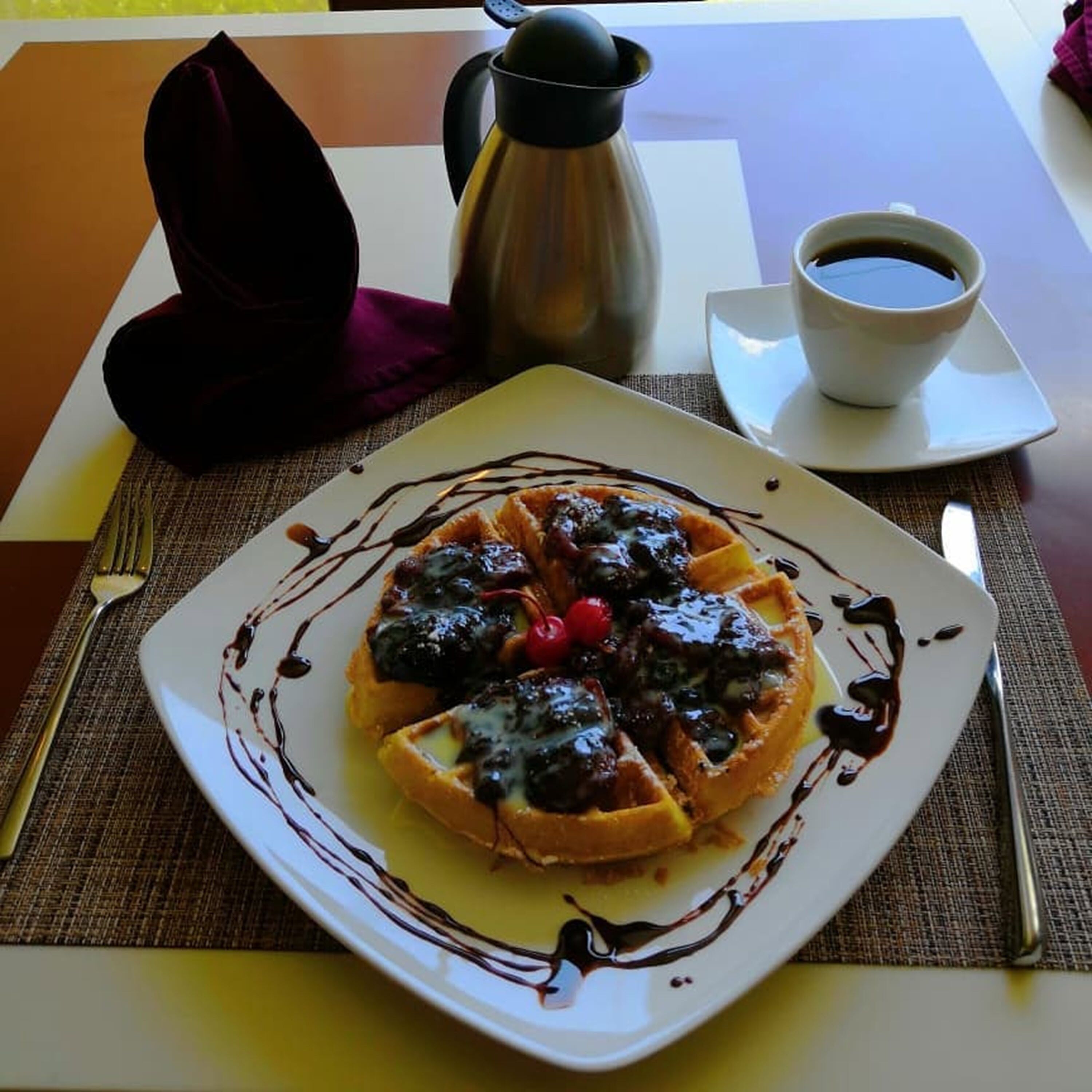Un piatto di waffle con mirtilli e sciroppo, una tazza di caffè e una thermos su un tavolo.
