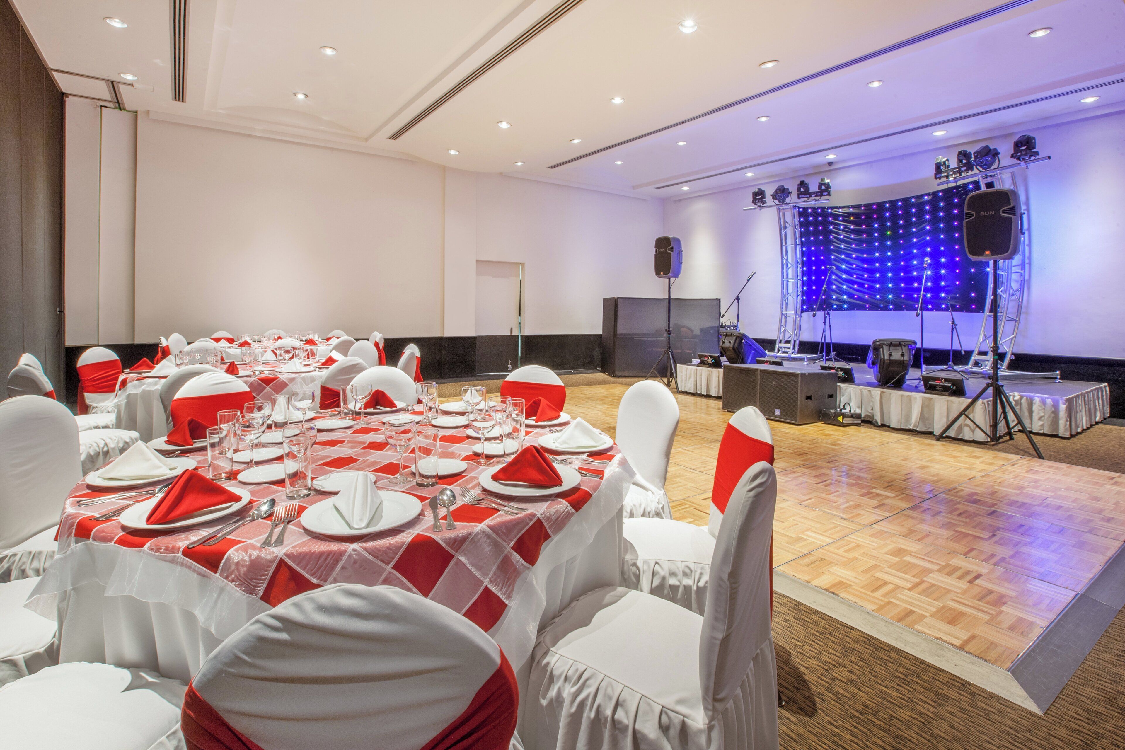 A banquet hall with round tables set for dining, a stage with lighting equipment, and a speaker system.