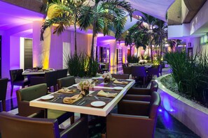 Comedor con una larga mesa dispuesta para una comida, rodeada de sillas, un arreglo de plantas tropicales y vistas a un pasillo.