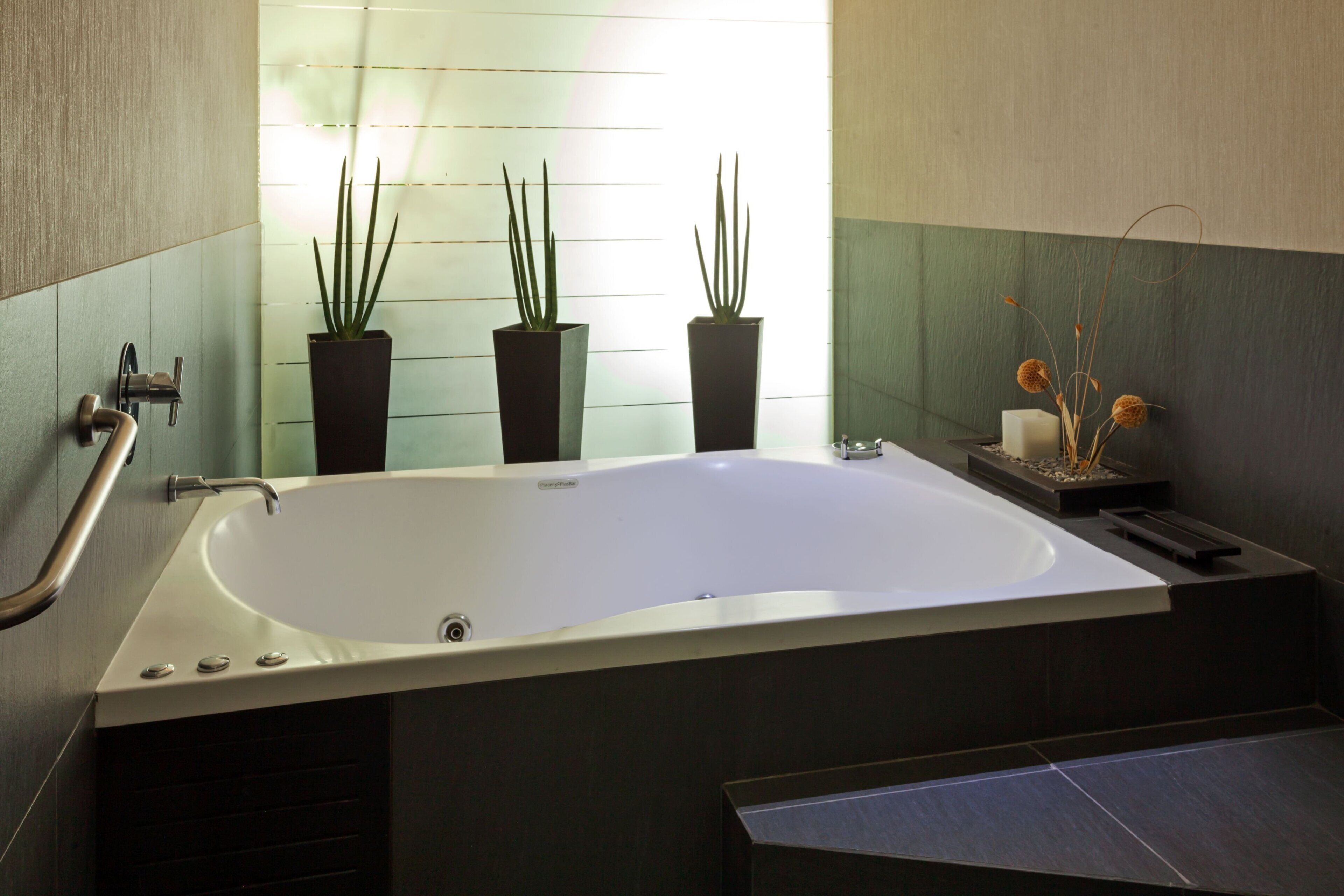 Un baño moderno con una bañera independiente, tres plantas en macetas y una mesita con una vela y objetos decorativos.