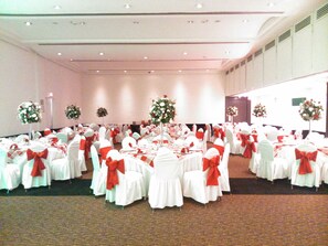 Un salón de banquetes con mesas redondas, manteles blancos y cintas rojas.
