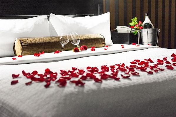 Una cama bien hecha con sábanas blancas, un edredón enrollado, dos copas de vino y pétalos de rosa esparcidos.
